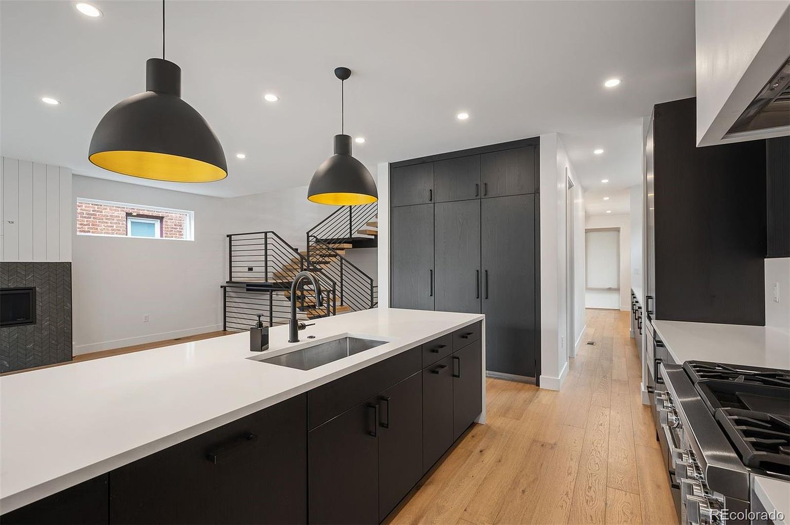 This is a modern kitchen with a large island featuring a white countertop and dark cabinetry. The kitchen includes stainless steel appliances and dark cabinets, with pendant lighting above the island. A staircase is visible in the background, and the overall design is sleek and contemporary.