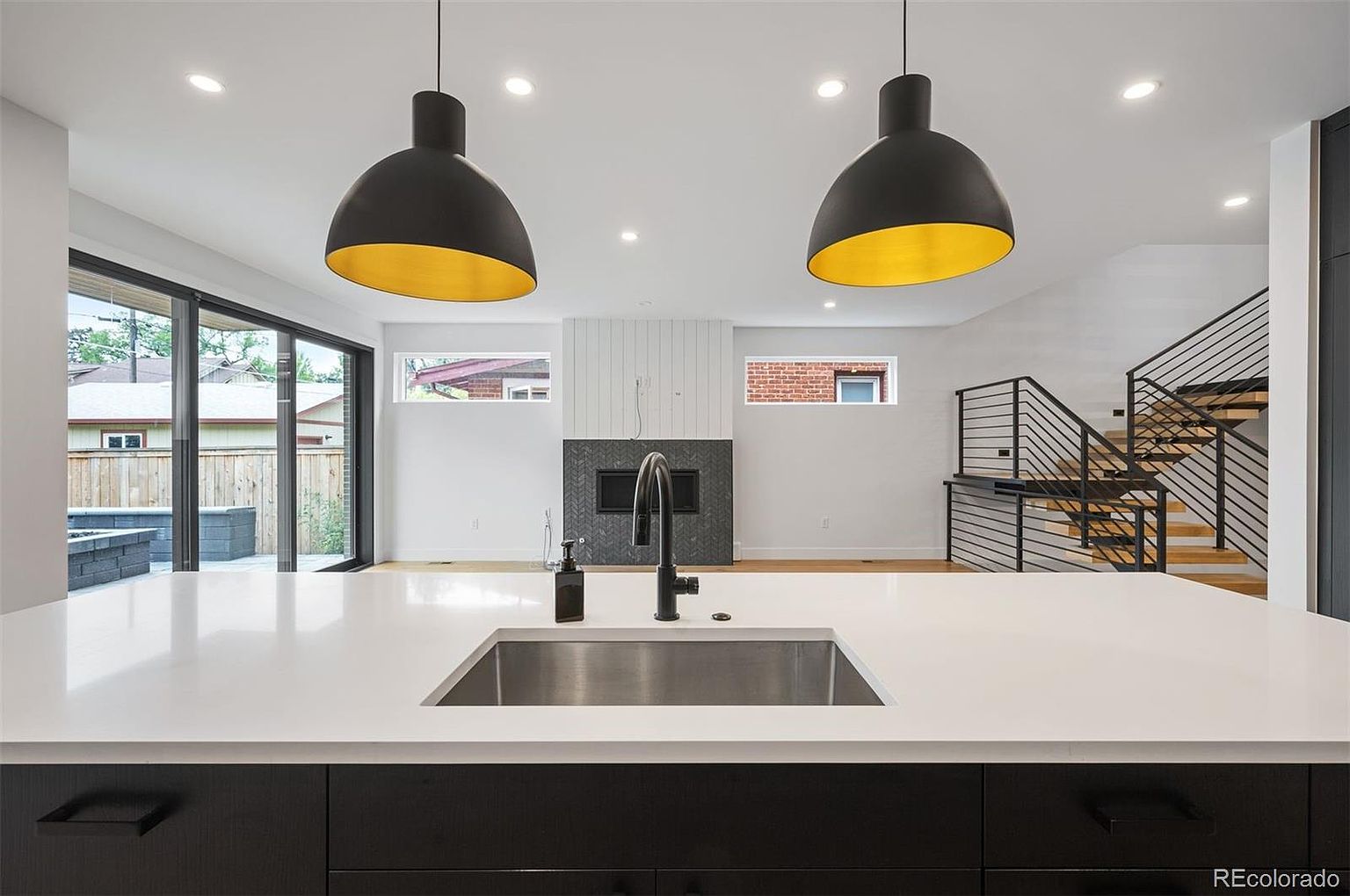 This interior shot showcases a modern kitchen with a large island featuring a stainless steel sink and black cabinetry. Two dome-shaped pendant lights with gold interiors hang above the island, illuminating the white countertops. The open-concept design flows into a living area with a fireplace and a staircase with black railings and wooden steps.