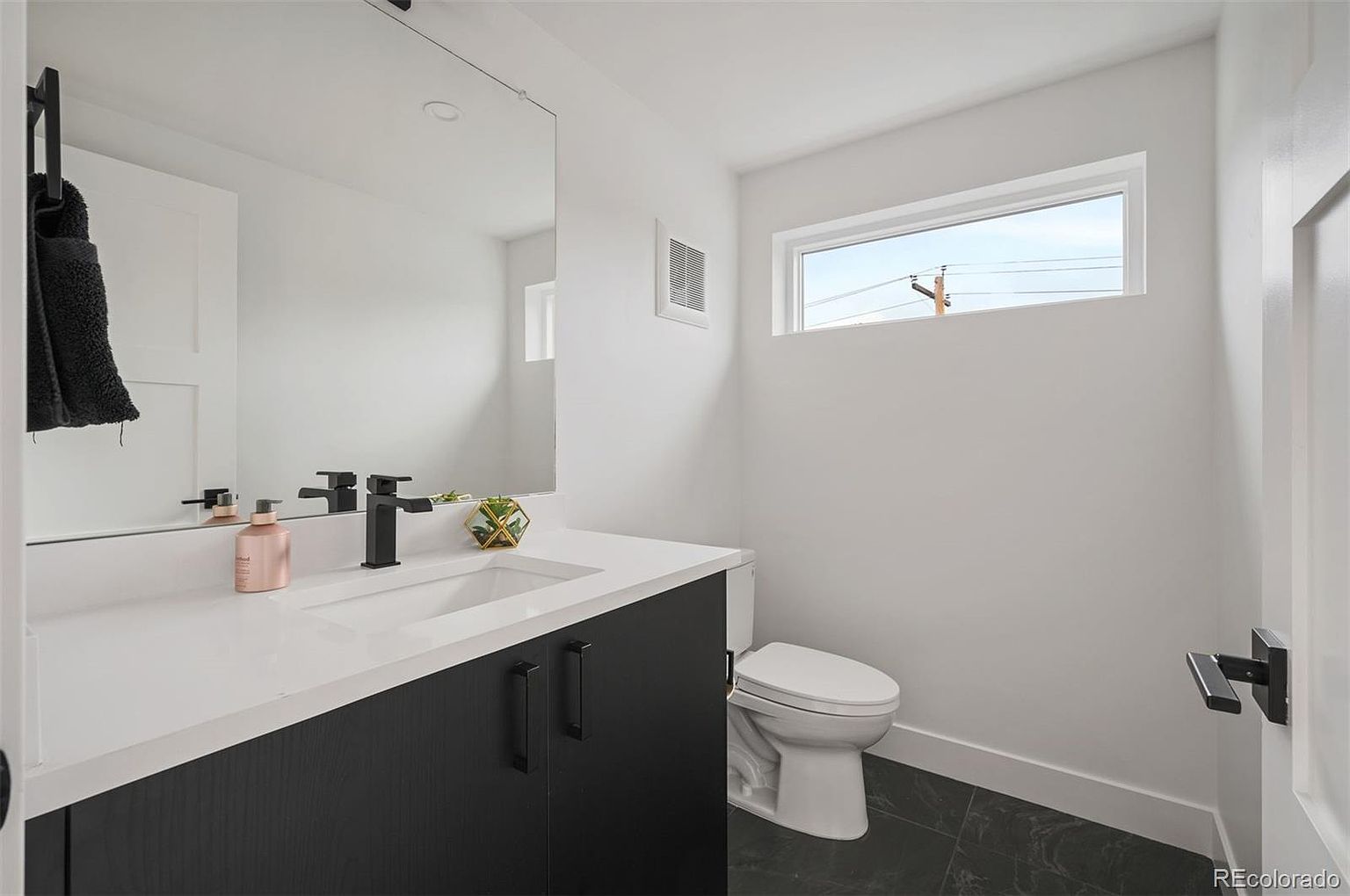 This is a modern guest bathroom featuring a sleek vanity with a white countertop and dark wood cabinetry. A large mirror reflects the space, enhancing the sense of openness. The room is well-lit with a window providing natural light, and the dark flooring adds a touch of sophistication.