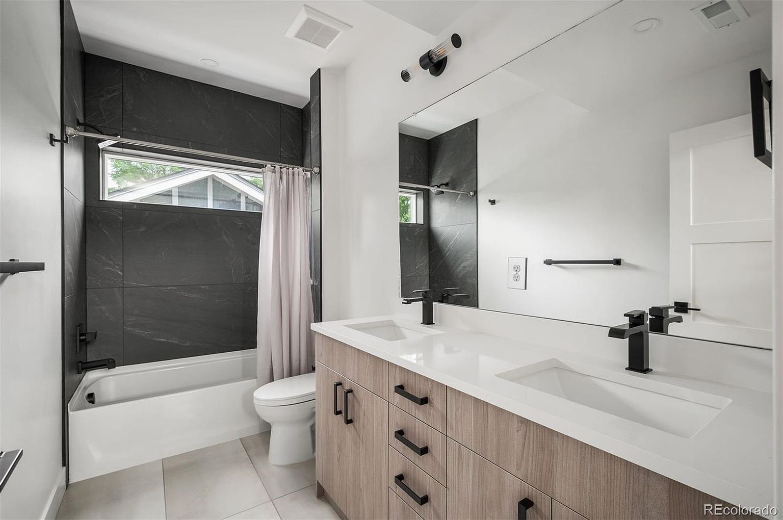This is a modern bathroom featuring a double vanity with light wood cabinets and black hardware. The countertops are white, and there are two black faucets. A large mirror reflects the shower/tub area, which has dark gray tile and a window. The floor is light gray tile, and the overall style is contemporary and clean.