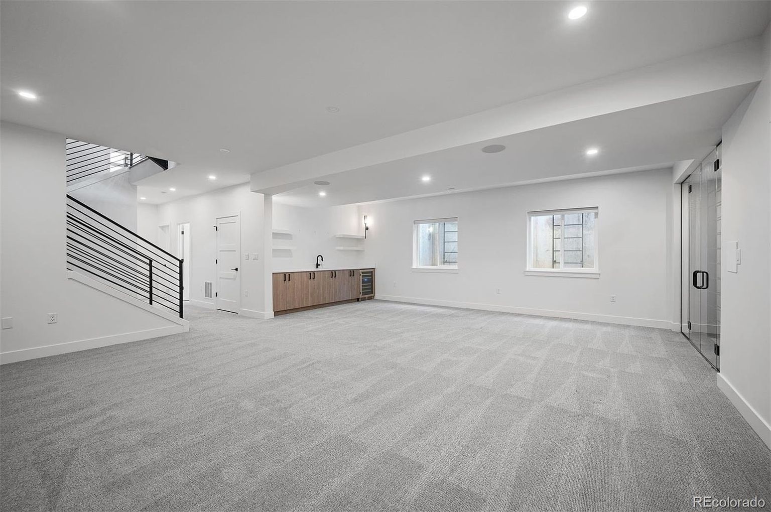 This is a spacious, modern basement featuring light gray carpeting and white walls. A staircase with black railings is visible on the left, and a wet bar with wooden cabinetry is situated along the back wall. Two windows provide natural light, and a glass door is on the right side of the room.