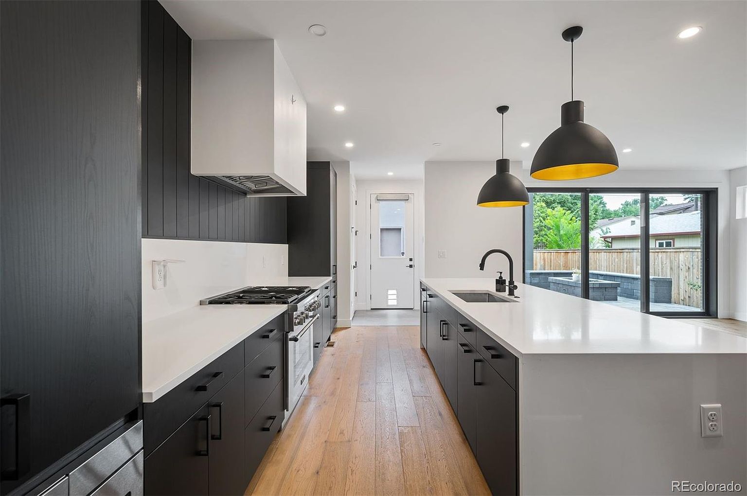This is a modern kitchen featuring dark cabinetry with sleek hardware and white countertops. The kitchen includes a gas range with a white hood and stainless steel appliances. Two pendant lights hang over the island, and a sliding glass door provides natural light and a view of the backyard.