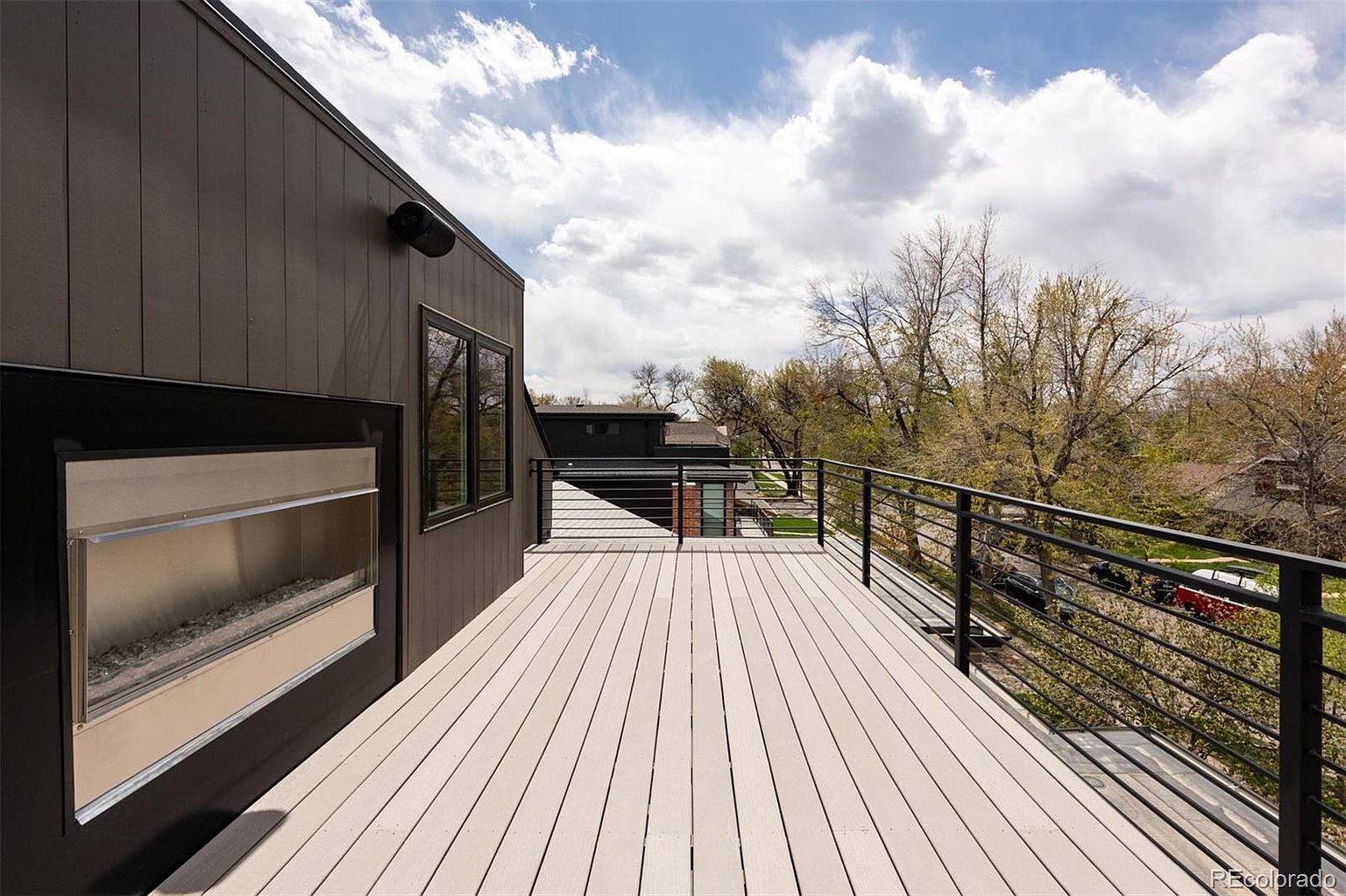 This image showcases a modern deck or balcony with light-colored wooden planks and a sleek black railing. A unique outdoor fireplace feature is built into the side of the building, adding a touch of luxury. The view from the deck includes trees and neighboring houses under a partly cloudy sky, suggesting a pleasant outdoor living space.