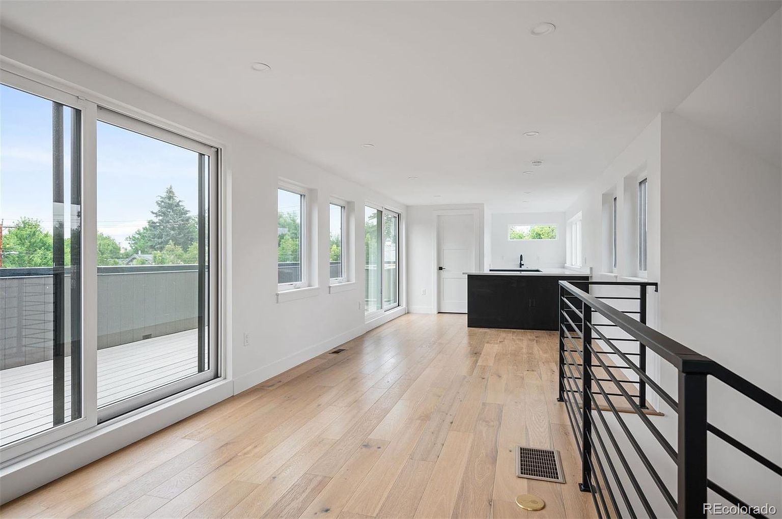 This is a bright and modern living room featuring light hardwood floors and white walls. Large windows provide ample natural light and views of the outdoors. A black kitchen island is visible in the background, and a black metal railing defines the staircase area, adding a contemporary touch to the space.