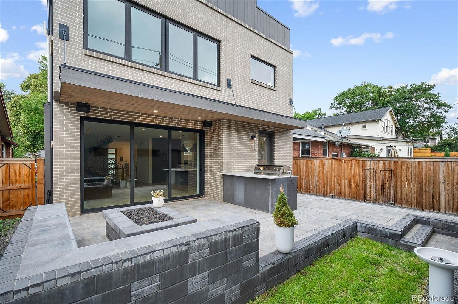 This image showcases a modern patio area with a built-in fire pit and outdoor kitchen featuring a grill. The patio is constructed with gray pavers and surrounded by a low brick wall, with a small patch of grass visible. Large sliding glass doors provide access to the interior of the home, creating a seamless indoor-outdoor living experience.