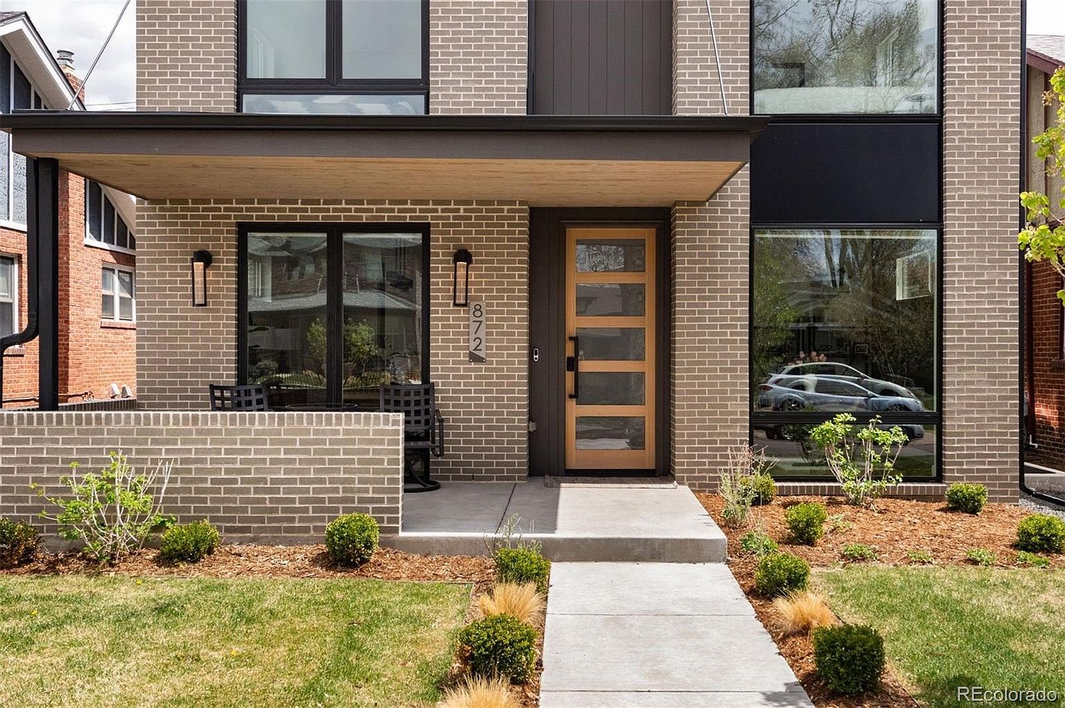 This is a front exterior view of a modern two-story home. The facade is primarily brick with dark trim around the windows and a light wood front door with glass panels. The landscaping includes a well-manicured lawn, mulch beds, and various shrubs, enhancing the curb appeal.