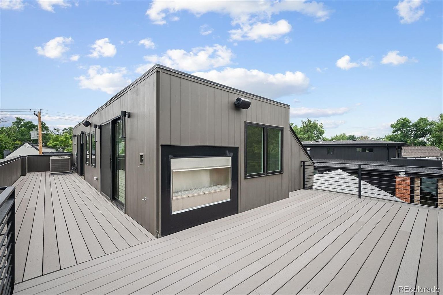This image showcases a modern rooftop deck featuring gray composite decking and sleek black metal railings. A contemporary gray-clad structure with black-framed windows and a unique fireplace feature adds architectural interest. The deck offers an open-air space for relaxation and entertainment, with views of the surrounding neighborhood.