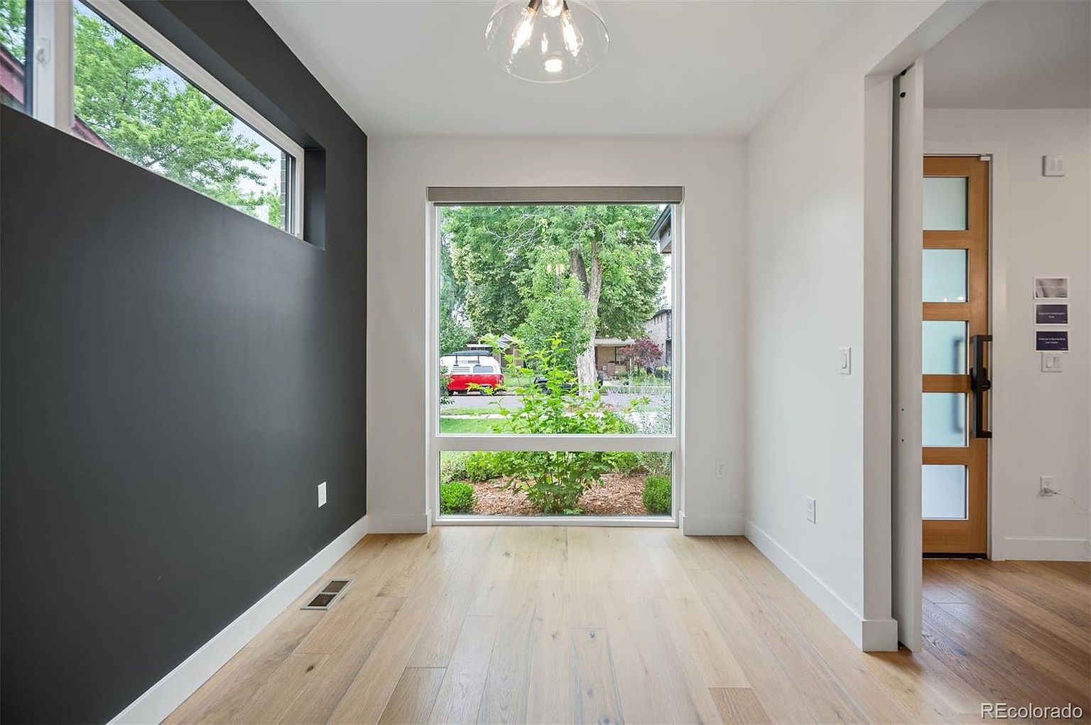 This is an interior shot of a modern dining room featuring light hardwood floors and a large window providing ample natural light. The room has a contemporary aesthetic with a dark accent wall and white trim, creating a clean and inviting space. A sliding door is visible on the right, adding to the room's functionality and style.