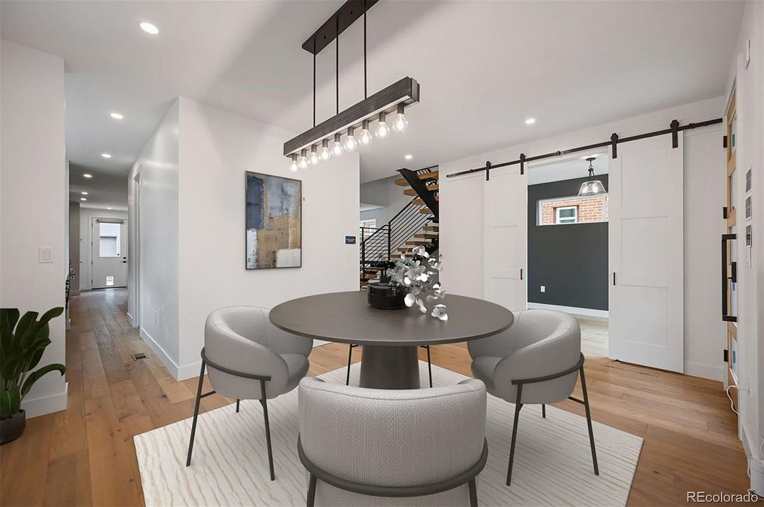 This interior shot showcases a modern dining room with a round table and four stylish chairs set on a light-colored rug. A contemporary chandelier hangs above the table, and artwork adorns the wall. The room features hardwood floors and white walls, creating a bright and inviting space, with sliding barn doors leading to another room.