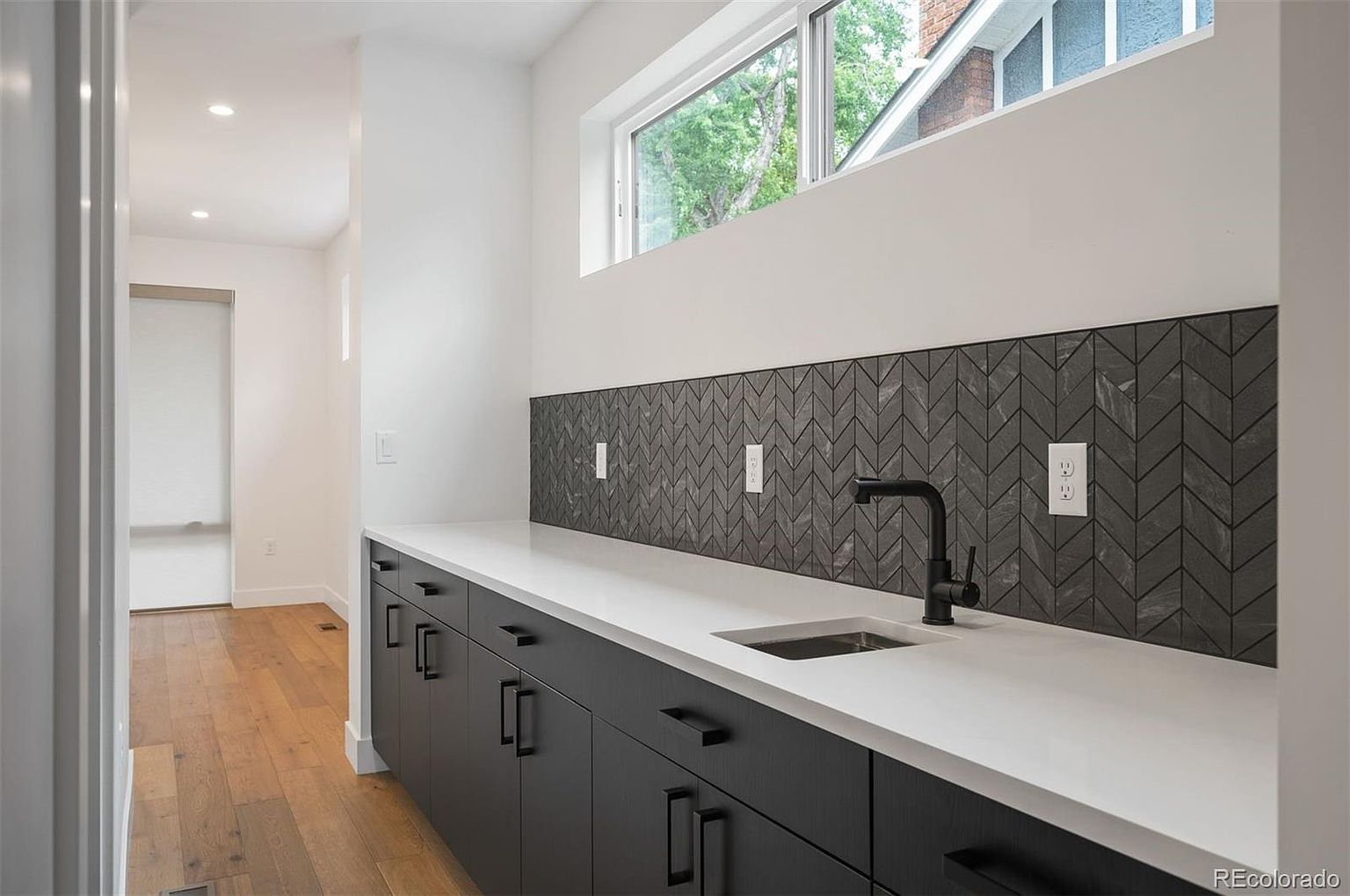 This interior shot showcases a modern kitchen area with sleek, dark cabinetry and a bright white countertop. A stylish black faucet complements the sink, set against a backdrop of patterned gray backsplash tiles. Natural light streams in through a window above, highlighting the clean lines and contemporary design.