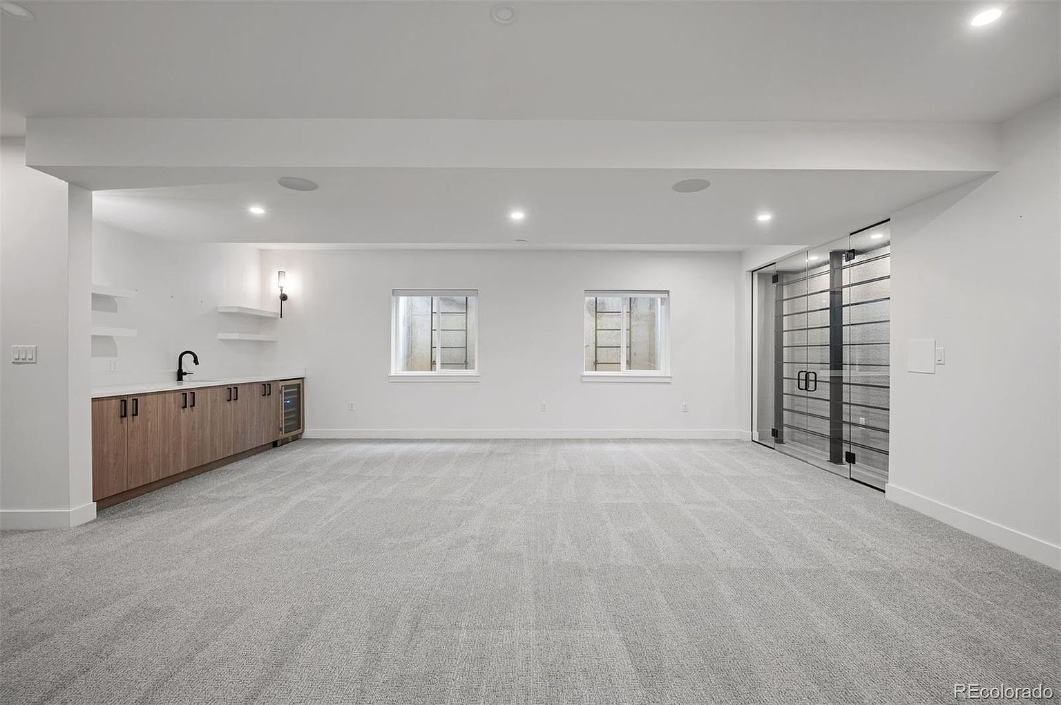 This is a spacious, modern basement featuring neutral gray carpeting and white walls. The room includes a wet bar with wood-tone cabinetry and open shelving, as well as two egress windows providing natural light. A glass-enclosed area, possibly a wine cellar or storage, adds a unique architectural element to the space.