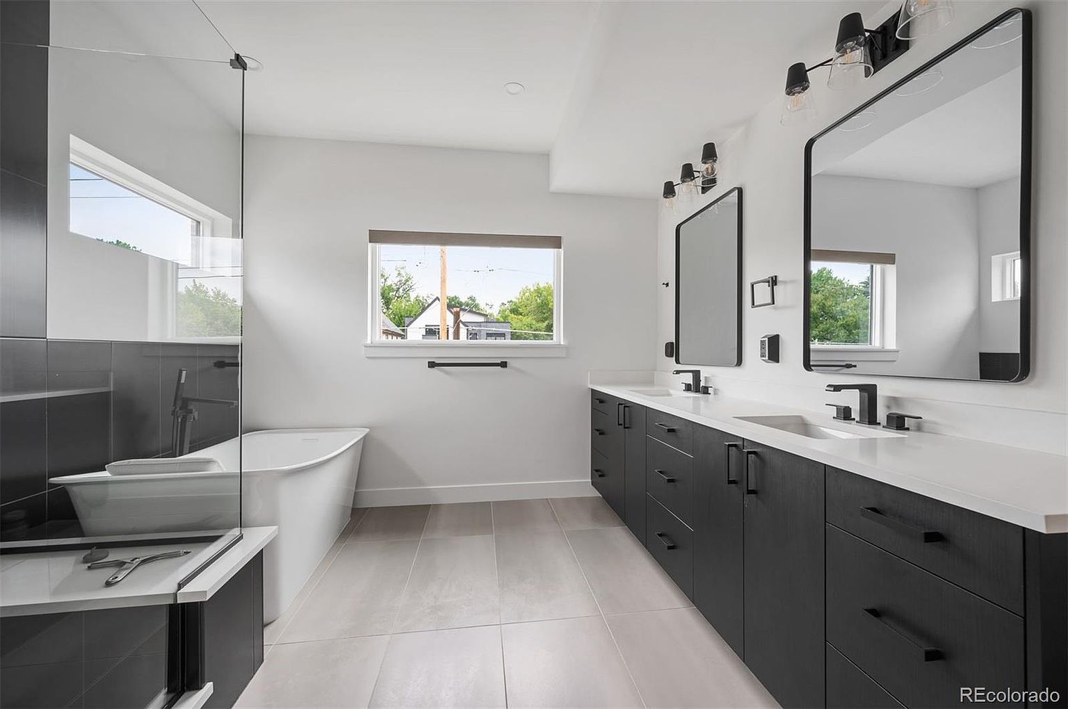 This is a modern primary bathroom featuring a double vanity with black cabinets and white countertops. Two rectangular mirrors hang above the sinks, illuminated by contemporary light fixtures. A freestanding bathtub is positioned near a window, and a glass-enclosed shower is visible to the left. The floor is tiled in a neutral tone, creating a clean and sophisticated aesthetic.