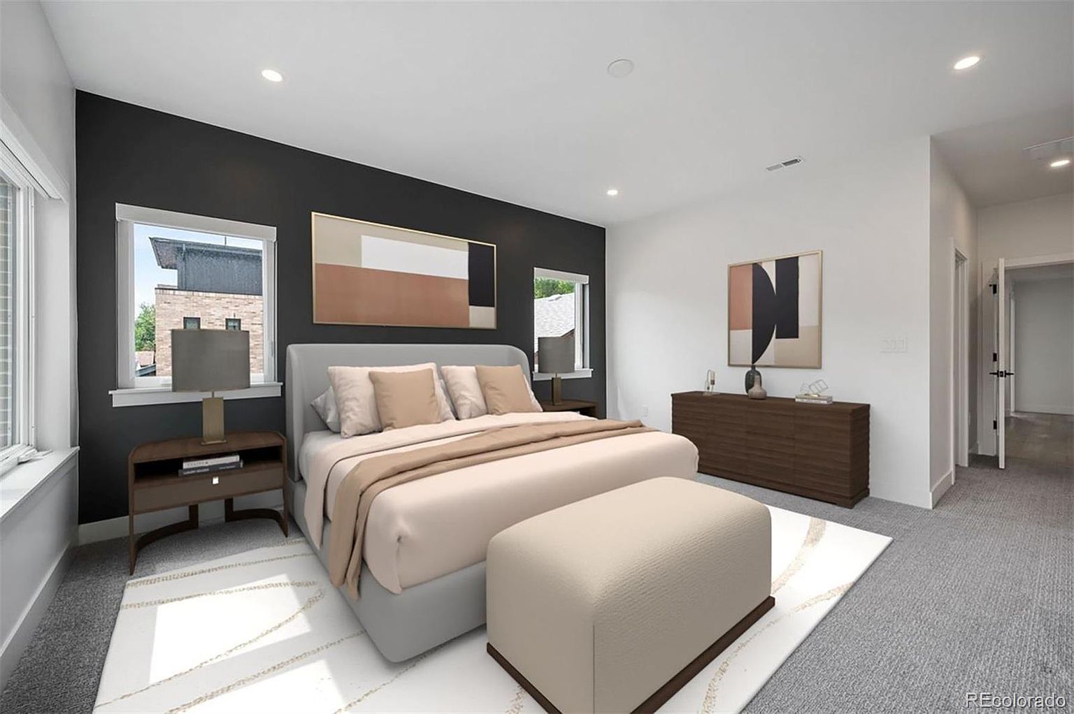 This is a staged primary bedroom featuring a gray upholstered bed with neutral bedding, flanked by a dark accent wall with modern artwork. The room is well-lit with natural light from two windows and recessed lighting, and includes a dark wood dresser and a beige ottoman at the foot of the bed. The overall impression is modern and serene.