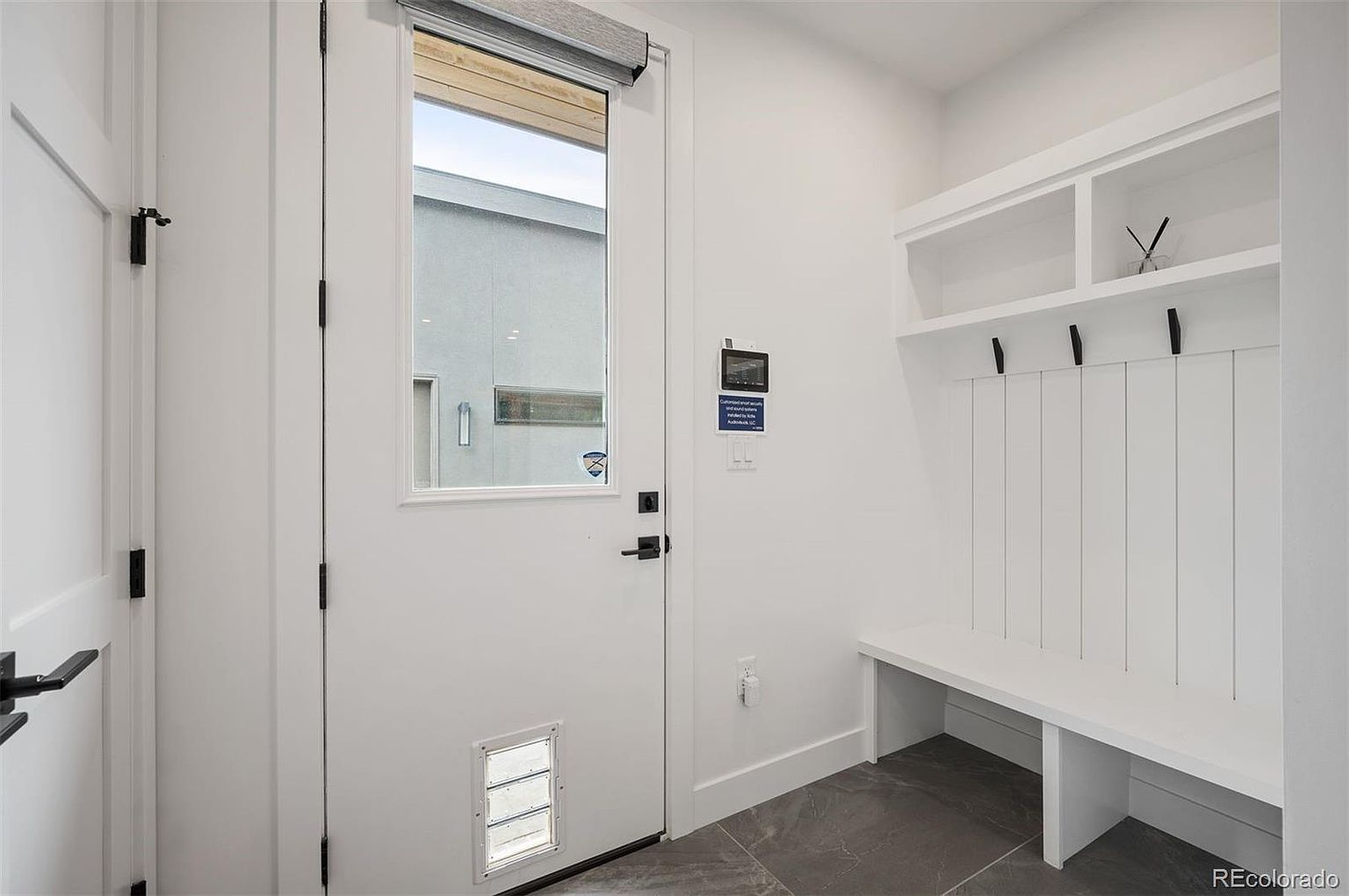 This interior shot showcases a modern hallway with a white door featuring a window and a pet door. To the right, a built-in bench and coat rack provide a functional mudroom area. The gray tile flooring and white walls create a clean and contemporary aesthetic.