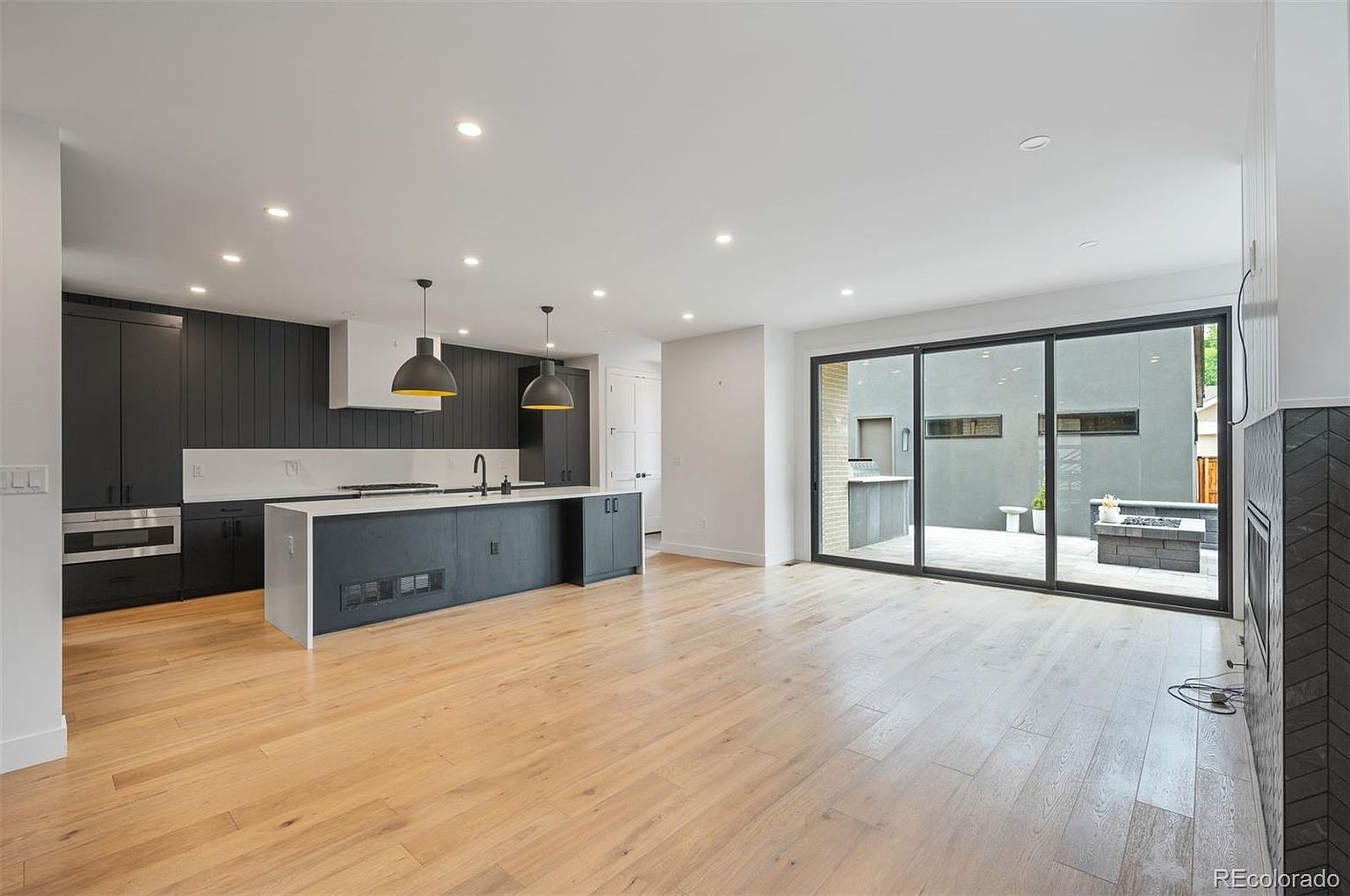This is a bright and modern kitchen and living area featuring dark cabinetry, a large island with a white countertop, and hardwood floors. The kitchen seamlessly transitions into a living space with a large sliding glass door leading to an outdoor patio. The perspective is a wide shot, showcasing the open-concept design and the connection between indoor and outdoor living spaces.