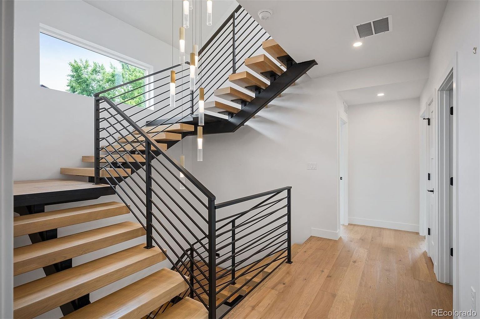 This interior shot showcases a modern hallway with a striking staircase. The staircase features wooden steps with black metal railings, complemented by contemporary pendant lighting. The hallway has light hardwood flooring and white walls, creating a bright and airy atmosphere, with doors leading to other rooms.