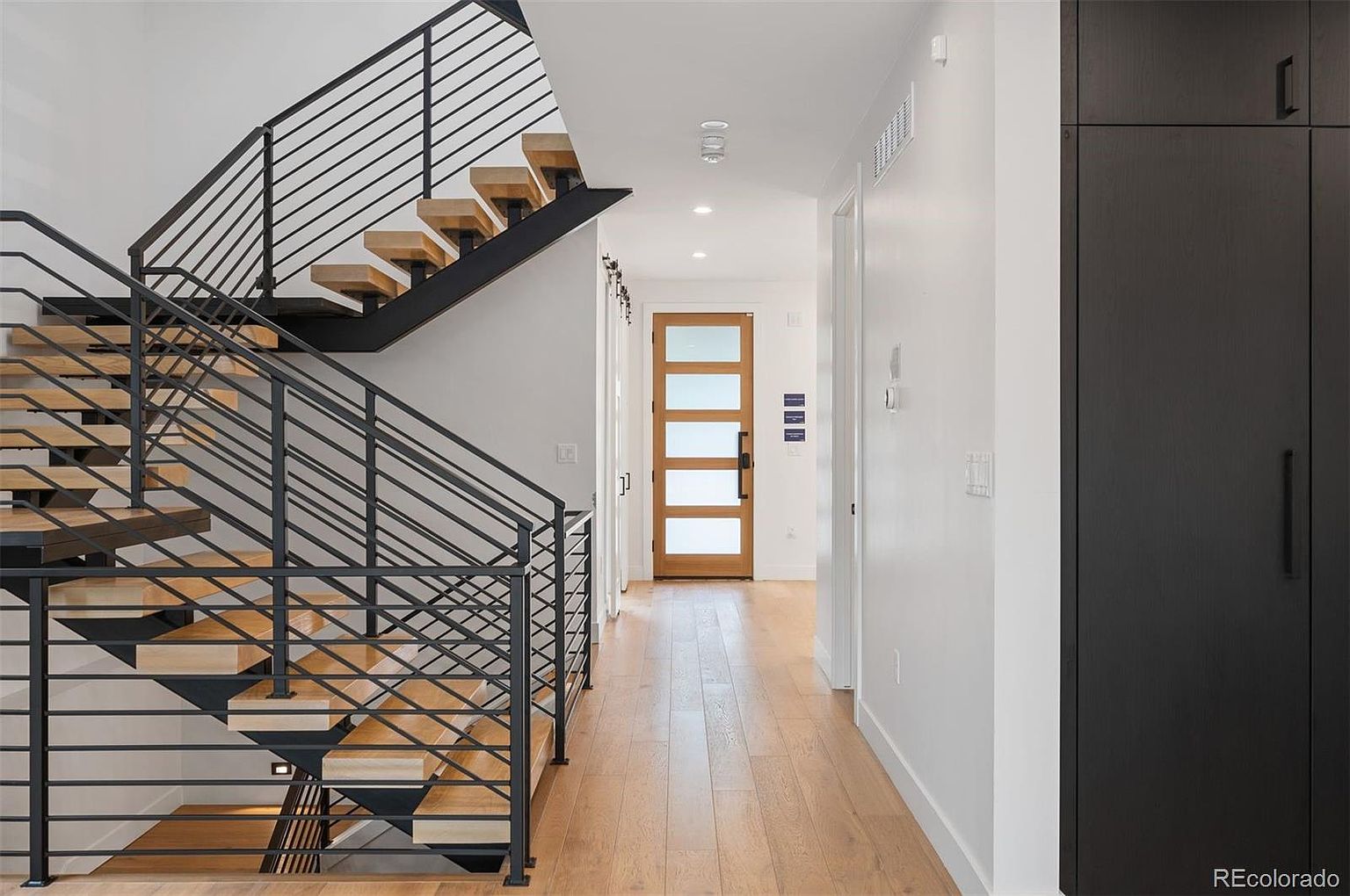 This interior shot showcases a modern hallway and staircase. The staircase features open wooden treads and a sleek black metal railing. The hallway leads to a light-filled room with a wooden door featuring frosted glass panels, while a dark cabinet stands against the wall on the right.