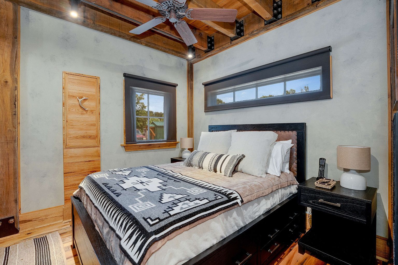 This is a cozy primary bedroom featuring a bed with a patterned blanket and several pillows. The room has exposed wooden beams on the ceiling and two windows that provide natural light. A wooden cabinet is built into the wall, and a nightstand with a lamp sits beside the bed, creating a warm and inviting atmosphere.