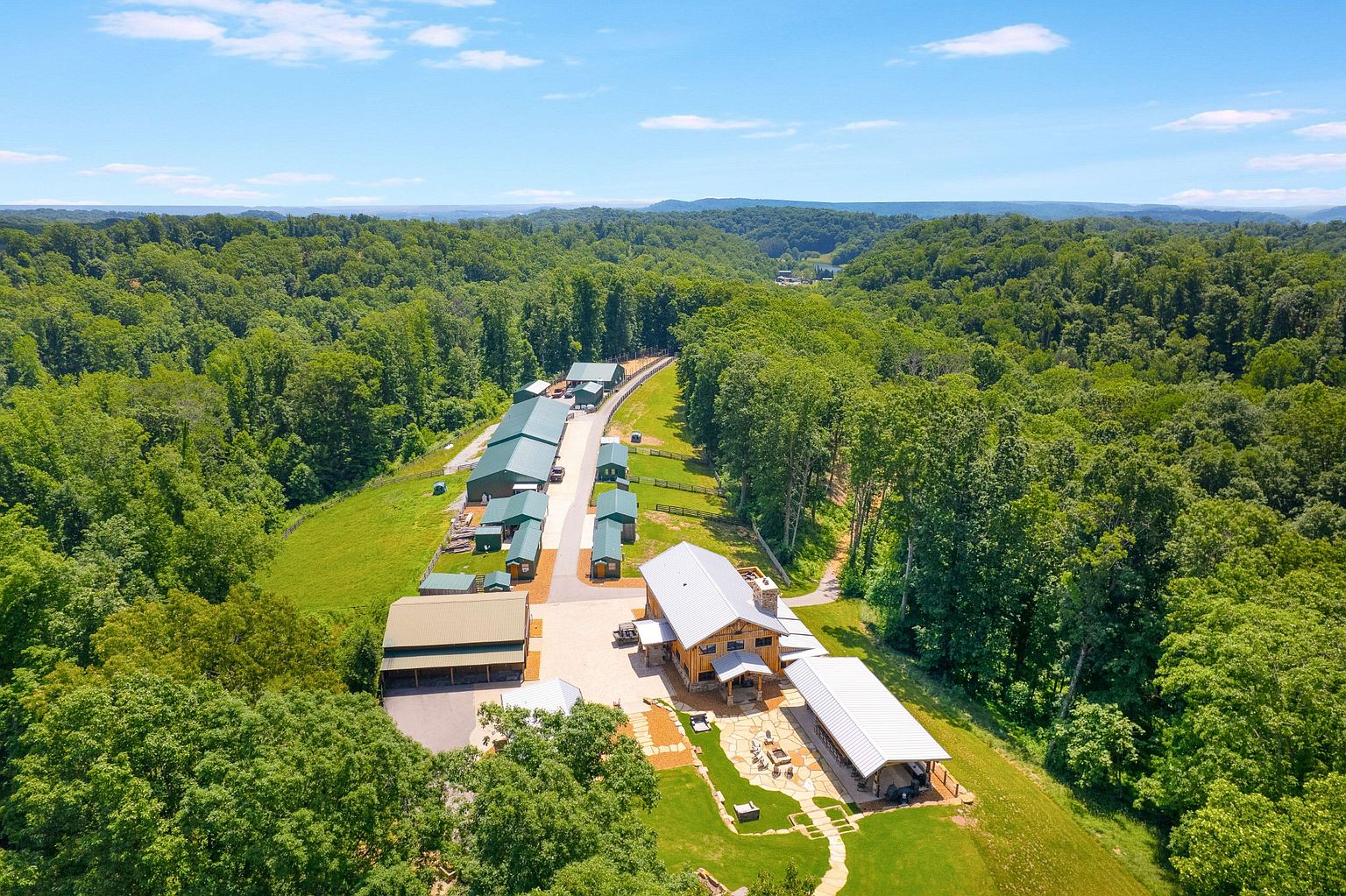 This aerial view showcases a sprawling property nestled amidst lush greenery. The property features multiple buildings with green roofs, a central wooden structure with a light-colored roof, and well-maintained landscaping with stone pathways and outdoor seating areas. The overall impression is one of a secluded and luxurious retreat.
