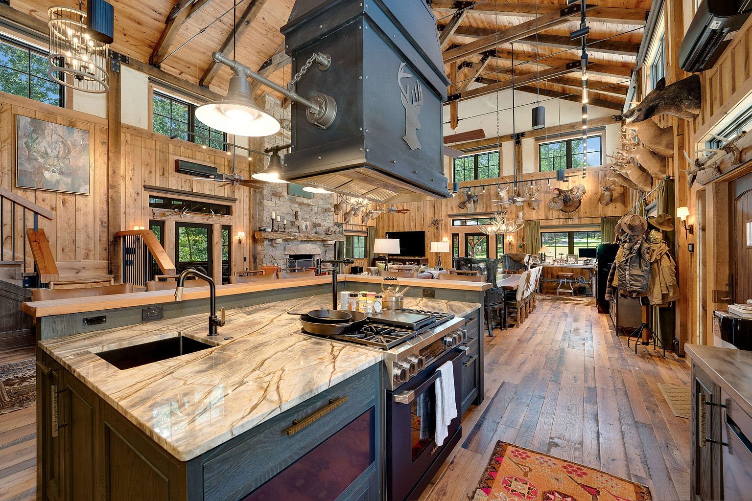 This is a spacious, rustic-style kitchen featuring wood-paneled walls and ceiling beams, a large kitchen island with a marble countertop, and a prominent range hood. The kitchen seamlessly blends into an open-concept living area with a stone fireplace and dining space, creating a warm and inviting atmosphere. Taxidermied animals adorn the walls, adding to the lodge-like ambiance.