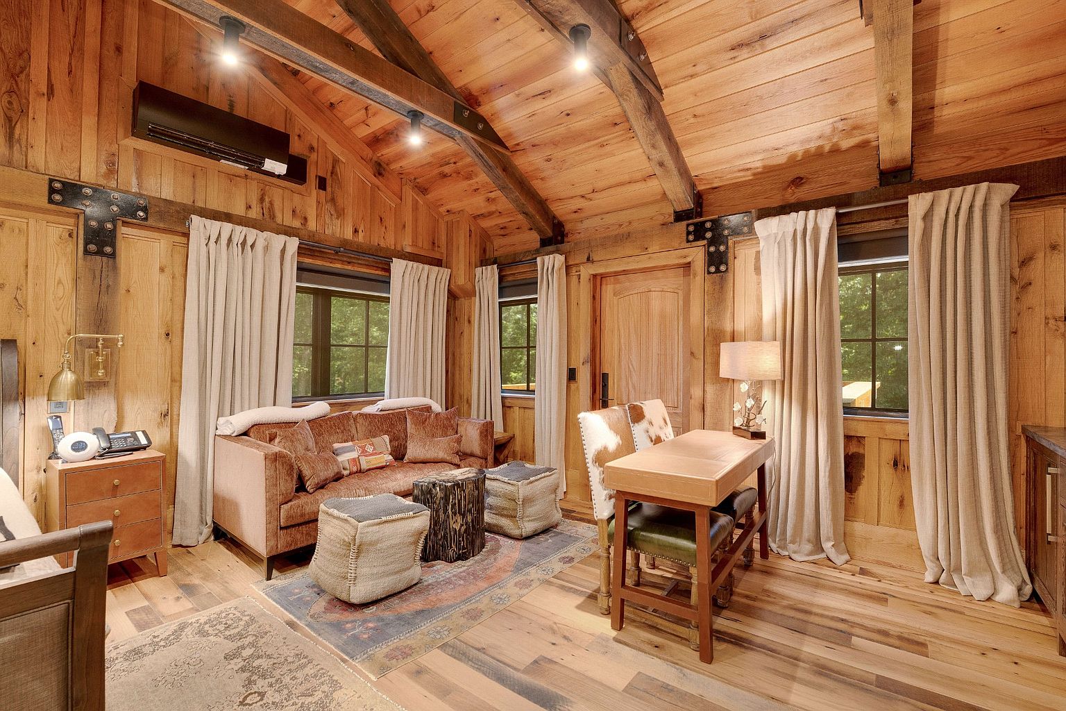 This is an interior shot of a cozy living room with wood-paneled walls and ceiling, accented by exposed beams. A comfortable brown sofa is complemented by unique stools and a patterned rug. A desk with chairs sits to the right, creating a functional workspace within the living area, and natural light filters through the windows, enhancing the warm and inviting atmosphere.