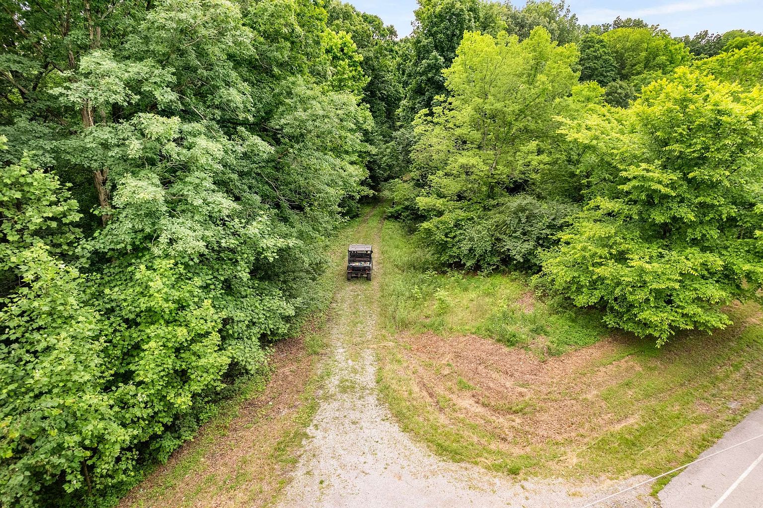 This aerial shot showcases a gravel driveway leading through a lush, green forest to a utility vehicle. The driveway is flanked by dense trees and foliage, creating a sense of seclusion and natural beauty. The perspective emphasizes the property's privacy and connection to nature, making it an appealing feature for potential buyers seeking a tranquil retreat.