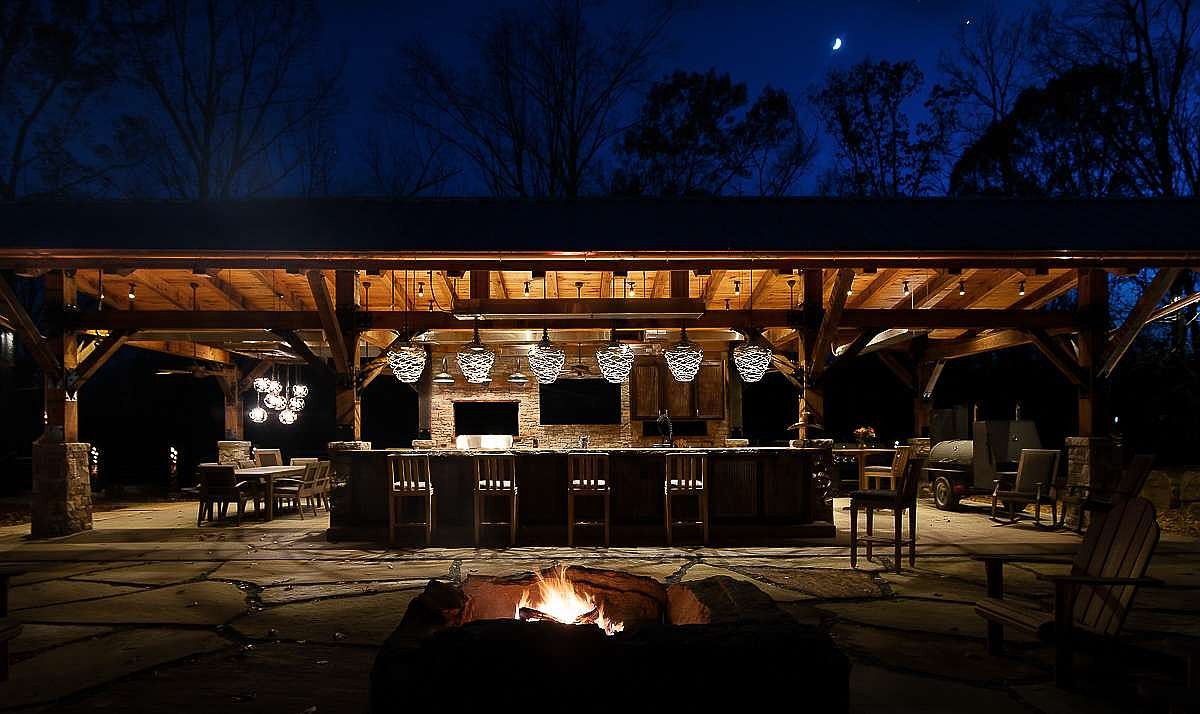 This image showcases a luxurious outdoor living space at night, featuring a covered patio with a built-in bar, complete with pendant lighting and bar seating. A stone fire pit in the foreground adds warmth and ambiance, while comfortable seating invites relaxation. The space is perfect for entertaining and enjoying the outdoors.