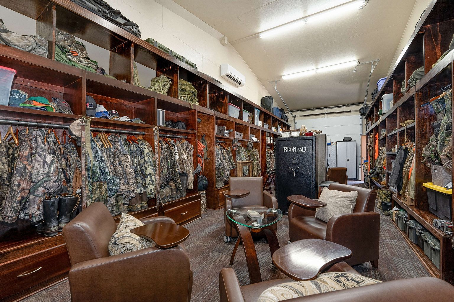 This is a well-organized walk-in closet featuring custom wooden shelving and hanging rods filled with camouflage clothing. Several leather armchairs with side tables are arranged in the center of the room, creating a lounge area. A large RedHead safe is visible in the background, adding a unique touch to the space.
