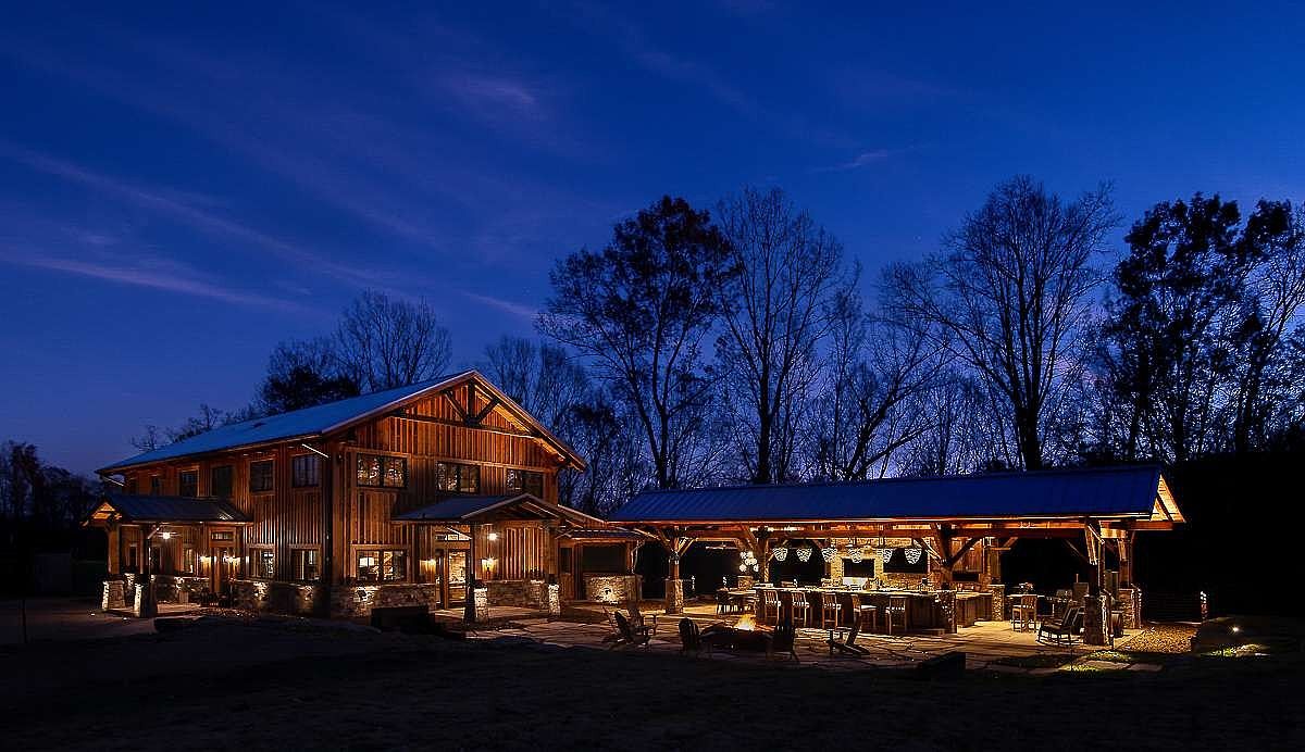 This image showcases the rear exterior of a stunning property at dusk, featuring a large wooden barn-style house and an adjacent outdoor entertainment area with a bar and seating. The structures are illuminated with warm lighting, highlighting the architectural details and creating an inviting atmosphere. The surrounding trees and dark blue sky add to the serene and luxurious ambiance, perfect for showcasing the property's outdoor living potential.