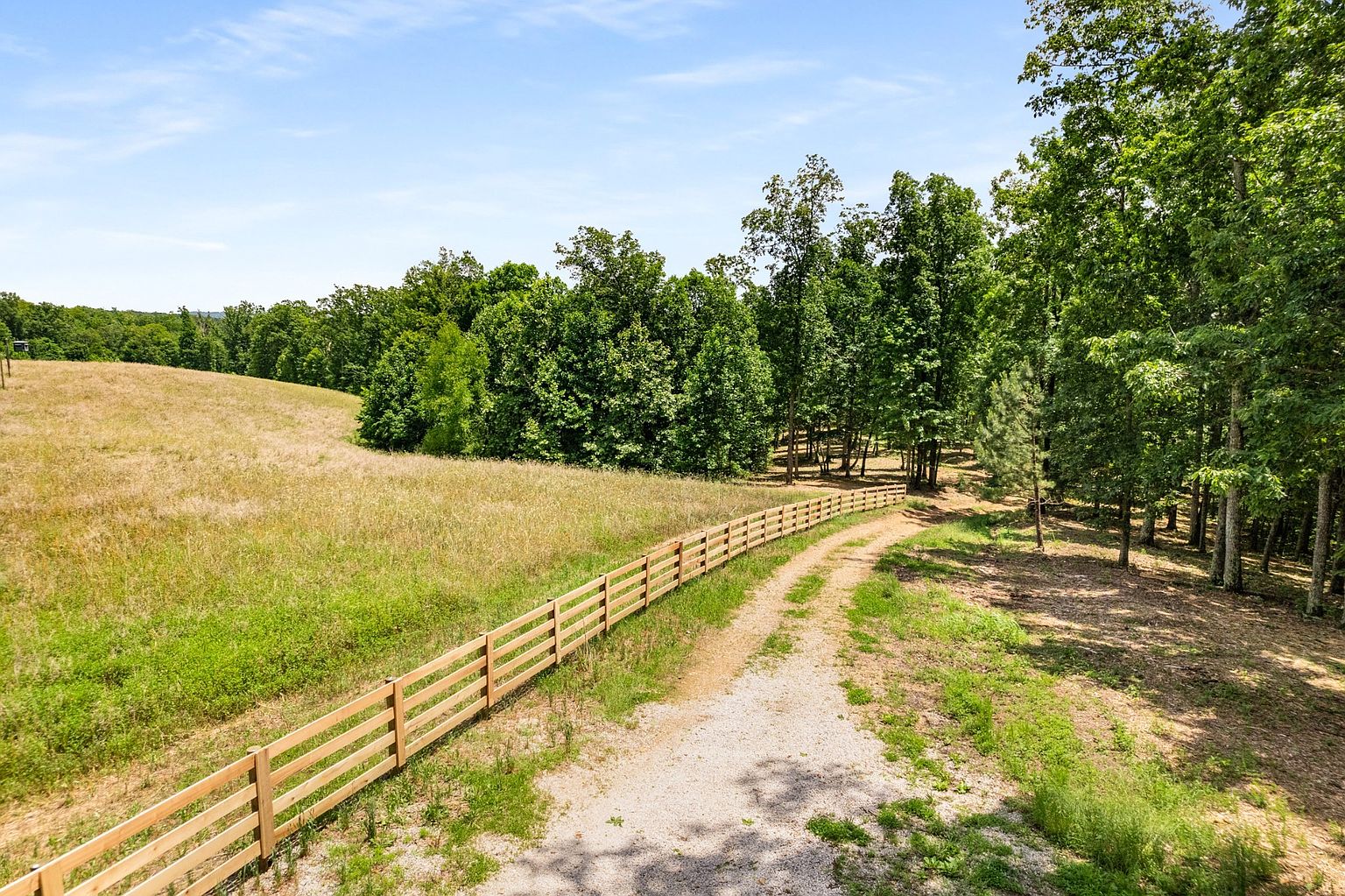 This aerial view showcases a sprawling property with a wooden fence bordering a grassy field and a dirt road leading into a wooded area. The landscape features a mix of open space and dense trees under a clear blue sky, creating a serene and private setting. The image highlights the property's size, natural beauty, and potential for various outdoor activities.