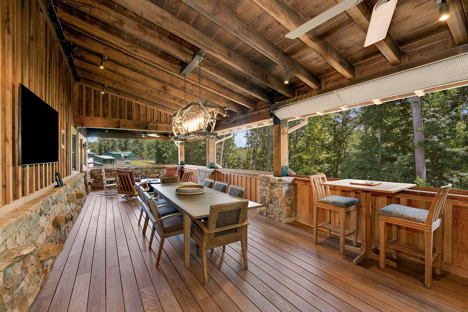 This image showcases a spacious outdoor patio or deck area, featuring a large wooden dining table with seating, a bar area with stools, and a stone-clad wall. The space is covered by a wooden ceiling with exposed beams and a unique chandelier, creating a rustic yet elegant atmosphere. The view opens to a lush green forest, enhancing the sense of privacy and tranquility.