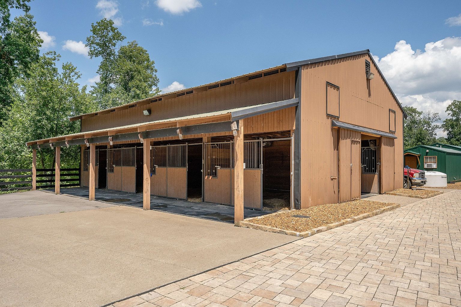 This image showcases the side view of a horse barn, featuring multiple stalls with wooden and metal gates. The barn has a brown exterior with a covered area supported by wooden posts, providing shelter for the stalls. A concrete area extends in front of the barn, transitioning to a brick-paved area on the right side, with a glimpse of a green outbuilding in the background.