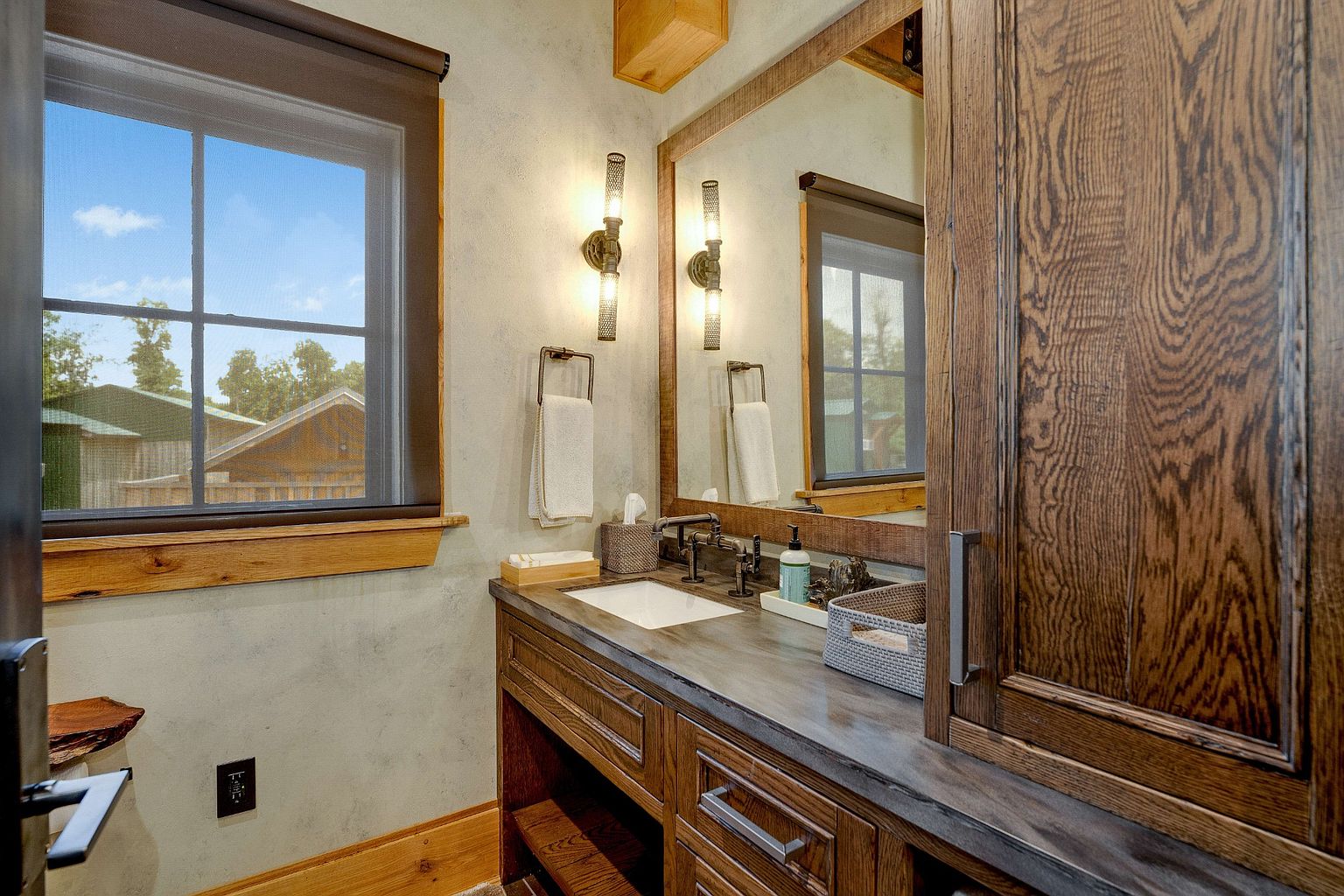 This is a well-lit bathroom featuring a rustic design. The vanity is made of dark wood with a light-colored countertop and a white sink. A framed mirror hangs above the sink, flanked by sconce lighting, and a window provides natural light. The overall impression is warm and inviting.