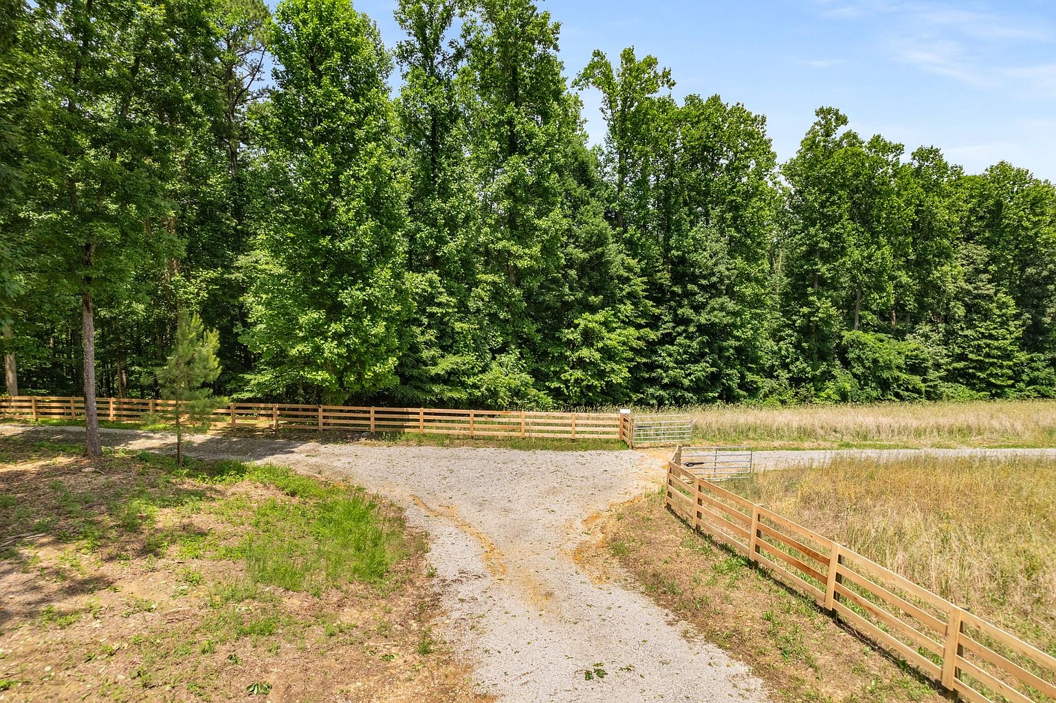 This image showcases a spacious yard or garden area, featuring a gravel driveway that splits into two directions, bordered by a wooden fence. Lush green trees line the background, creating a natural and private setting. The overall impression is one of a well-maintained and inviting outdoor space, perfect for a rural property.
