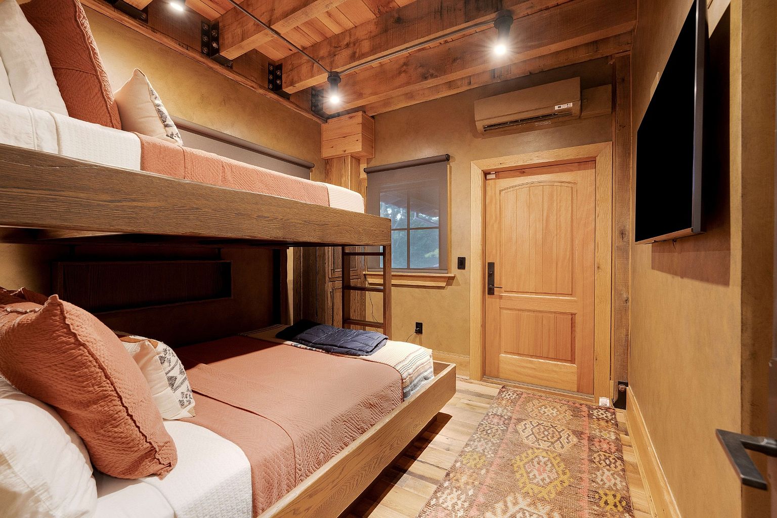 This is an interior shot of a rustic bedroom featuring custom-built bunk beds with wooden frames and cozy bedding. The room has a warm, inviting ambiance with wooden floors, textured walls, and a decorative rug. A window provides natural light, and a flat-screen TV is mounted on the wall, enhancing the room's appeal as a comfortable and stylish guest space.