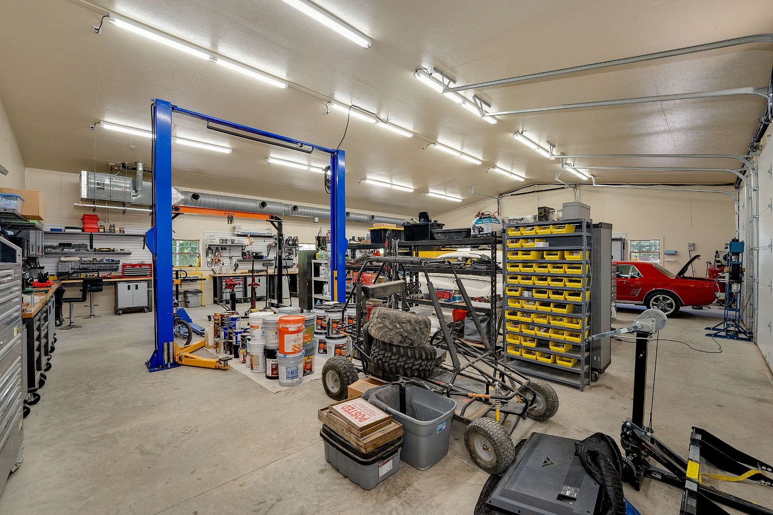 This is a spacious garage featuring a car lift, various tools, and storage solutions. The concrete floor and ample lighting create a functional workspace. A classic red car is parked in the background, adding a touch of character to the scene.
