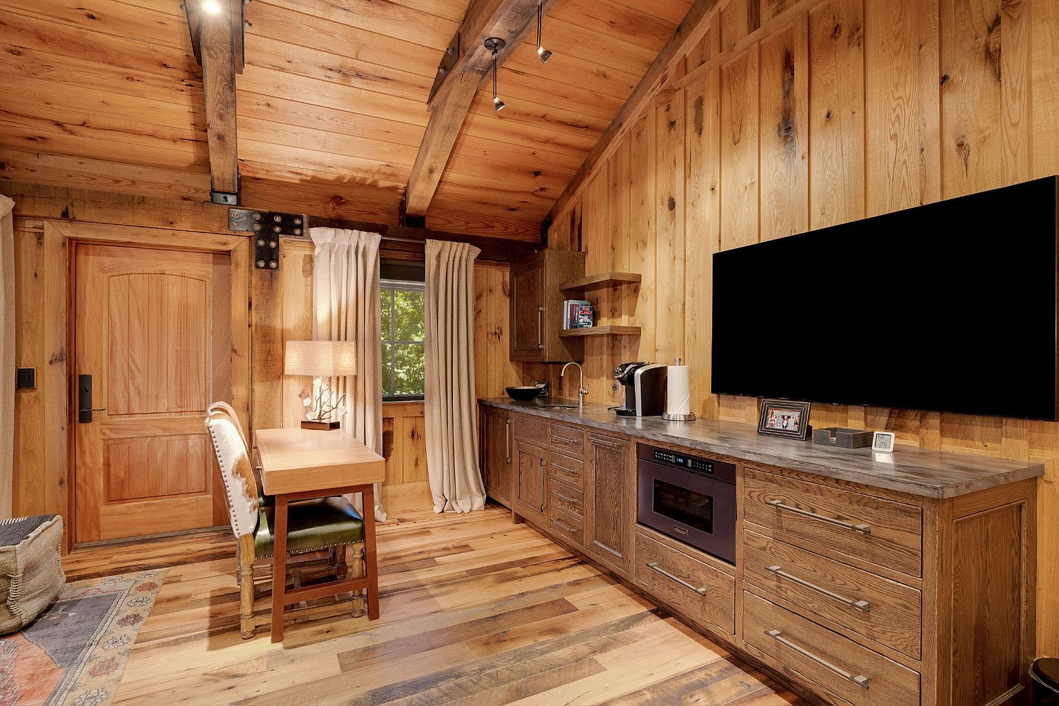 This interior shot showcases a rustic office space with wood-paneled walls and ceiling, creating a warm and inviting atmosphere. A wooden desk and chair are positioned near a window, while a built-in counter with a microwave and coffee maker provides convenience. A large television is mounted on the wall, adding a modern touch to the room.
