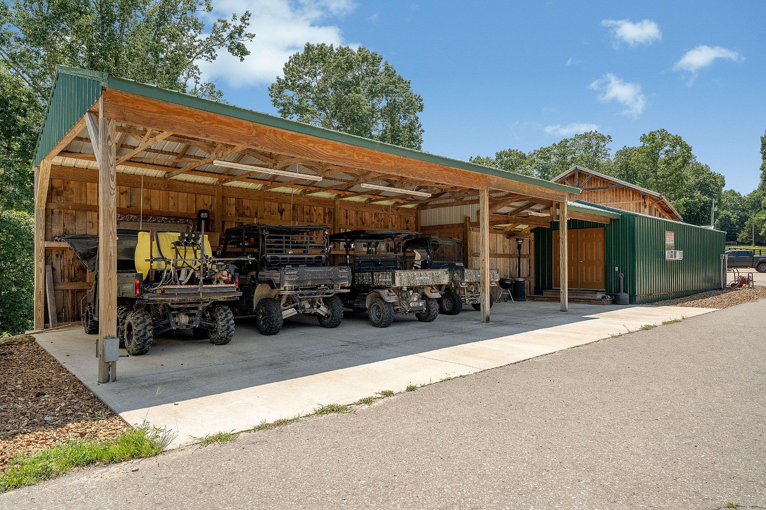 The image showcases an exterior view of a large, open-sided garage or storage structure, housing several all-terrain vehicles. The structure features a wooden frame with a metal roof, and the vehicles are parked on a concrete slab. The overall impression is one of functionality and ample storage space, suitable for a rural or recreational property.