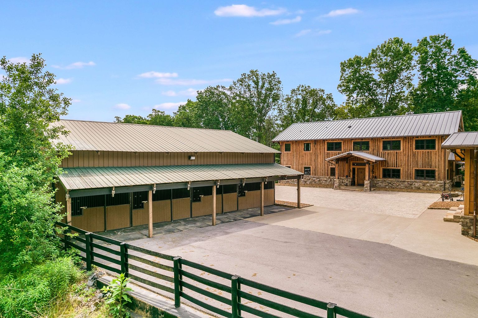 This aerial view showcases a well-maintained equestrian property featuring a stable with multiple stalls and a large barn-style house. The property includes a spacious concrete driveway and is surrounded by lush greenery and mature trees. The overall impression is one of a luxurious and functional rural estate.