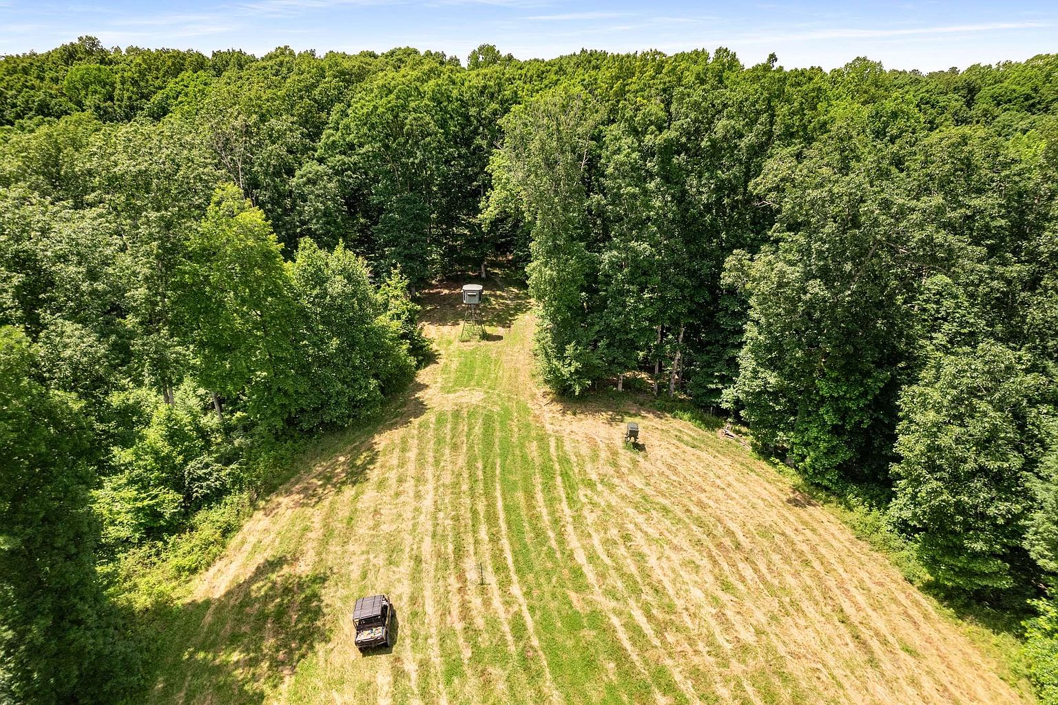 This aerial shot showcases a vast property featuring a meticulously maintained field bordered by dense, lush trees. A vehicle is parked at the edge of the field, and a hunting blind is visible further up the path, suggesting recreational opportunities. The image conveys a sense of privacy and spaciousness, ideal for a buyer seeking a secluded retreat.