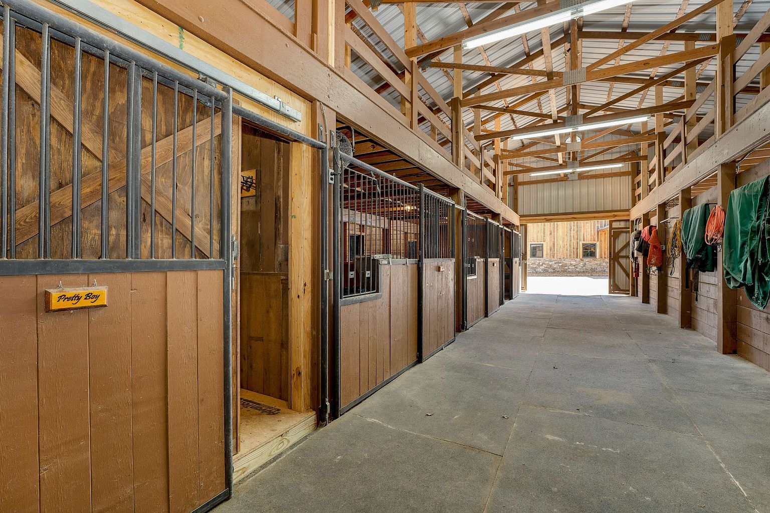 This image showcases the interior of a horse barn, featuring a row of stalls with metal bars and wooden panels. The barn has a high ceiling with exposed wooden beams and metal roofing, illuminated by fluorescent lights. The concrete floor extends down the hallway, leading to an open doorway at the end, creating a sense of depth and spaciousness.