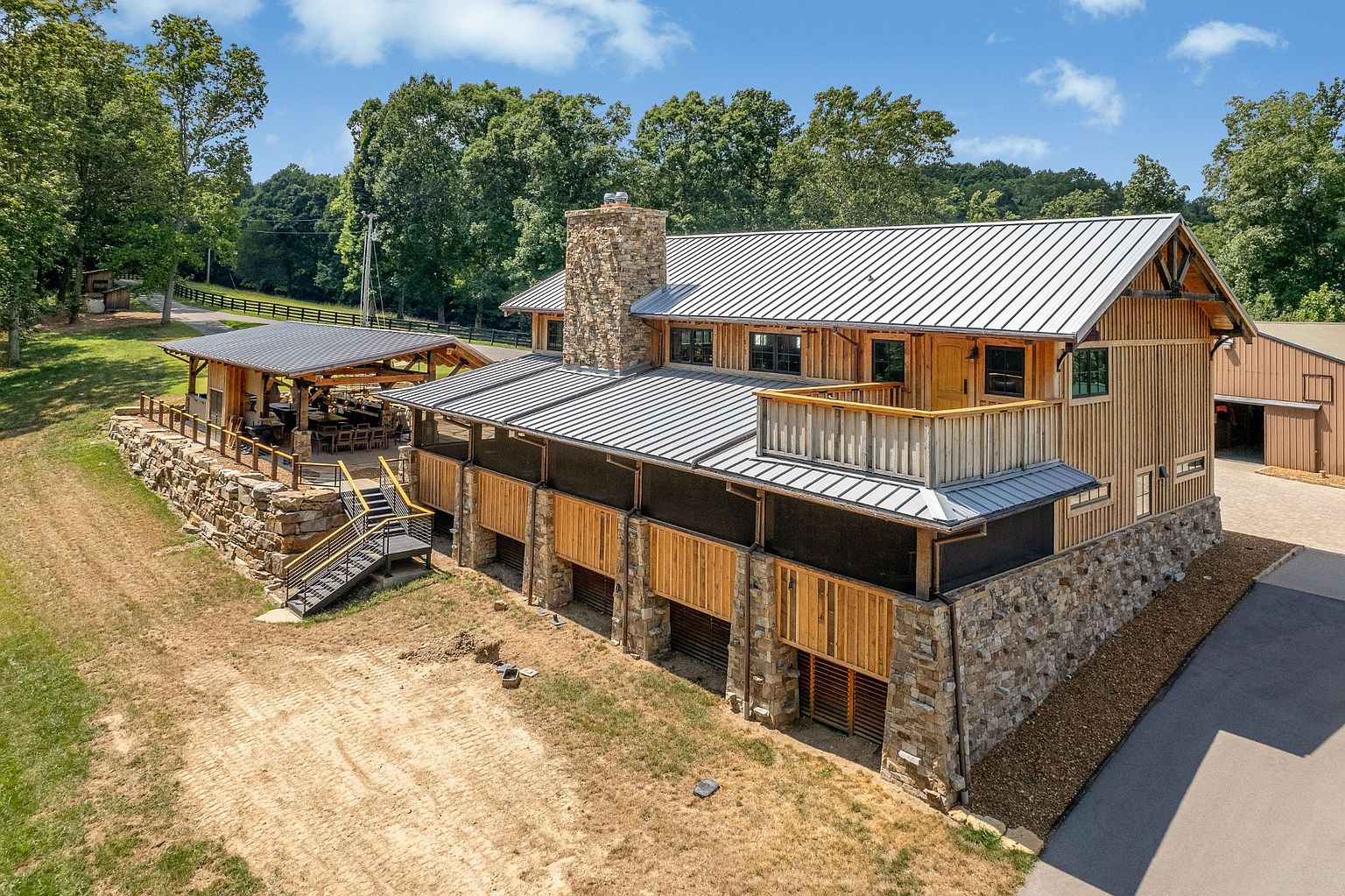 This aerial view showcases a stunning multi-level wooden and stone building with a metal roof, complemented by an adjacent outdoor structure with a dining area. The property features a well-maintained lawn and landscaping, highlighting the expansive grounds and architectural details. A paved driveway leads to the building, suggesting easy access and ample parking.