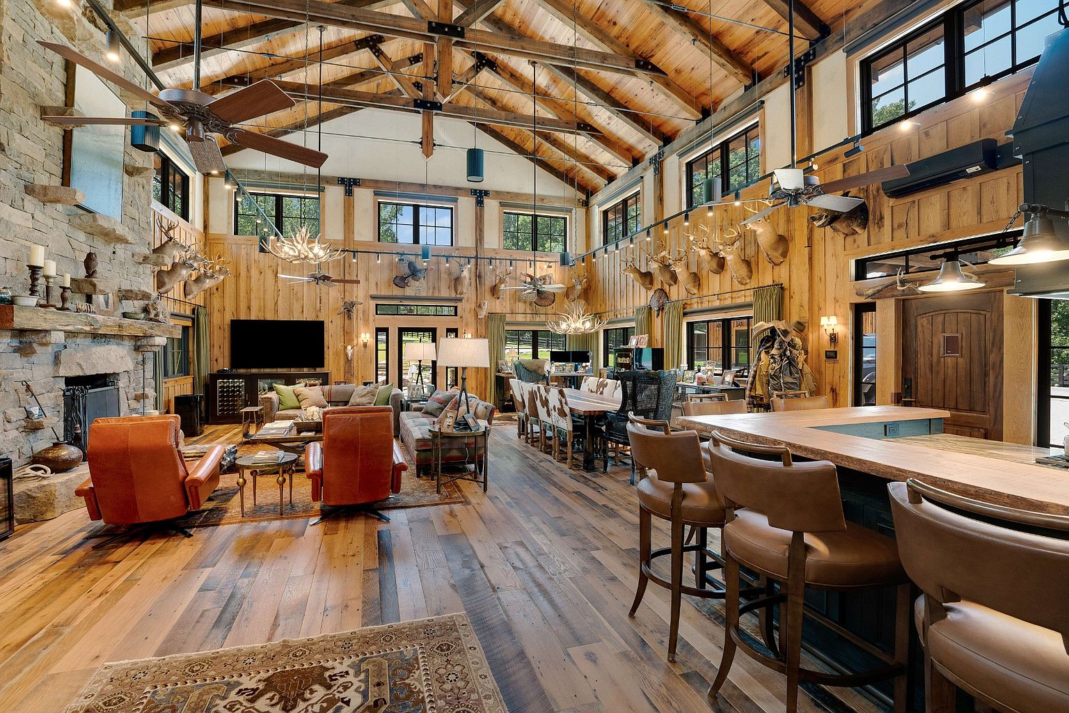 This grand living room features a soaring wood-paneled ceiling with exposed beams, a stone fireplace, and an open floor plan that seamlessly integrates the living, dining, and kitchen areas. The space is decorated with rustic-chic furnishings, including leather armchairs, comfortable sofas, and unique antler chandeliers, creating a warm and inviting atmosphere. The room is filled with natural light from the many windows.