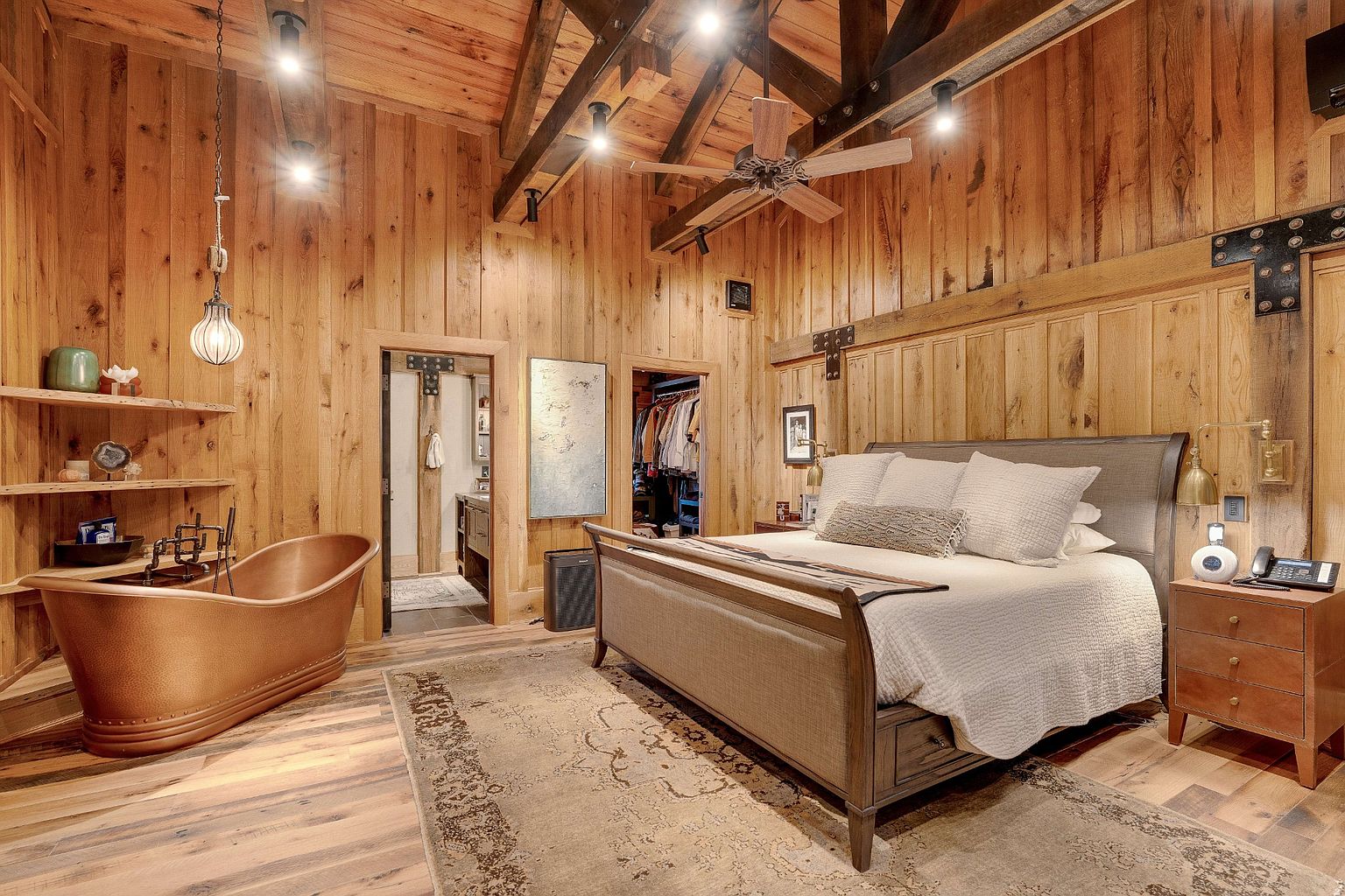 This is a primary bedroom featuring wood paneled walls and ceiling beams, creating a rustic and luxurious ambiance. The room includes a large bed with neutral bedding, a copper bathtub, and access to a walk-in closet and bathroom. The overall impression is warm and inviting, highlighting the unique architectural details and high-end finishes.