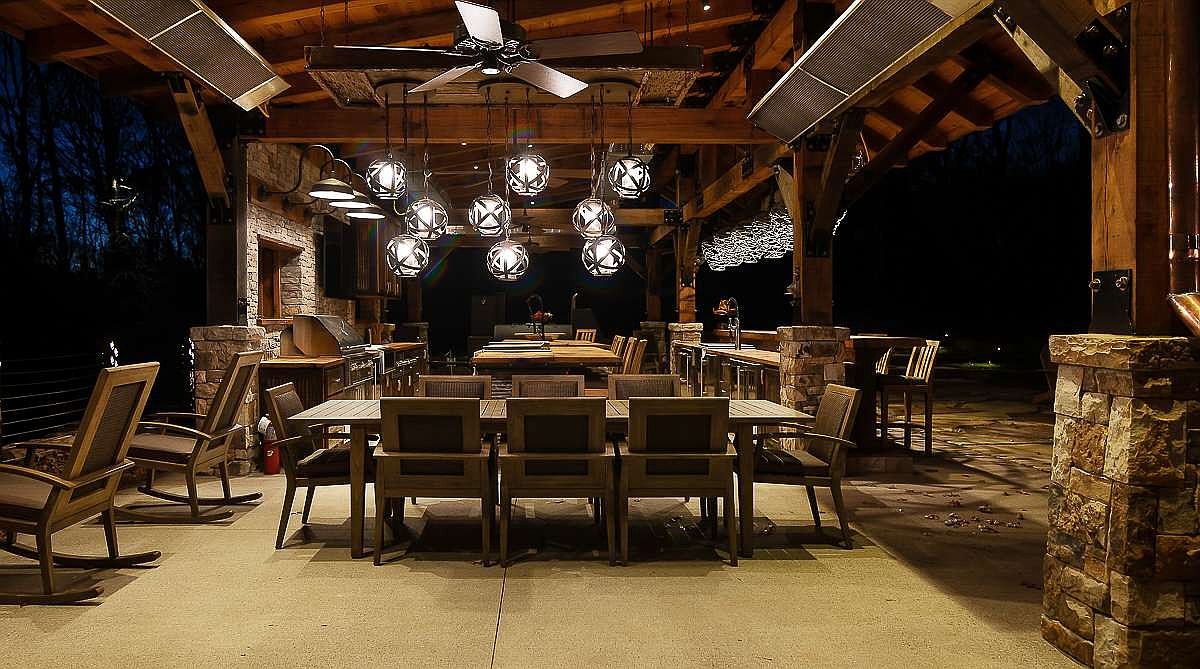 This image showcases a beautifully designed outdoor patio area, featuring a large wooden pergola with a ceiling fan and decorative lighting. The space is furnished with a dining table and chairs, as well as rocking chairs, creating a comfortable and inviting atmosphere for outdoor dining and relaxation. Stone pillars and a built-in grill area add to the luxurious feel of the space.