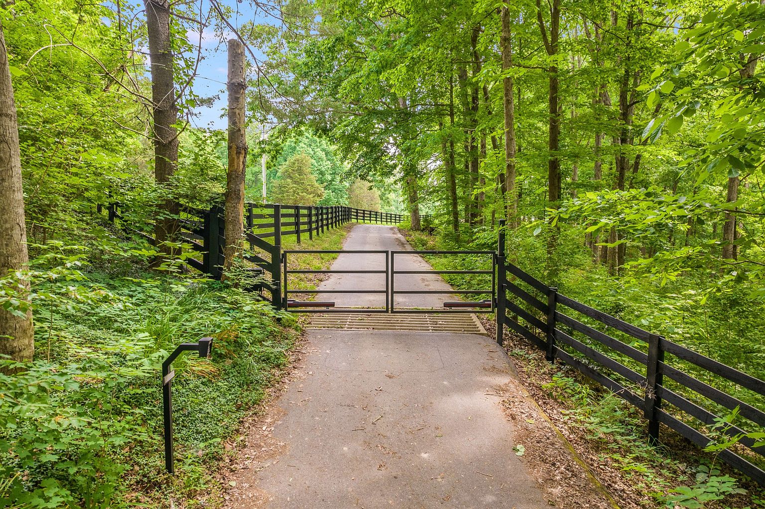 The image showcases a gated entryway leading to a property, framed by lush greenery and mature trees. A black wooden fence lines the driveway, adding a touch of rustic elegance. The asphalt driveway leads into the distance, creating a sense of privacy and seclusion.