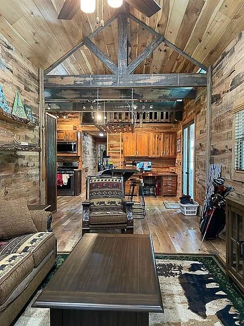 This interior shot showcases a rustic living room with wood-paneled walls and ceiling, creating a warm and inviting atmosphere. The room features a comfortable sofa, an armchair, and a wooden coffee table, complemented by a patterned rug. The open layout leads into the kitchen area, enhancing the sense of spaciousness and connectivity within the home.
