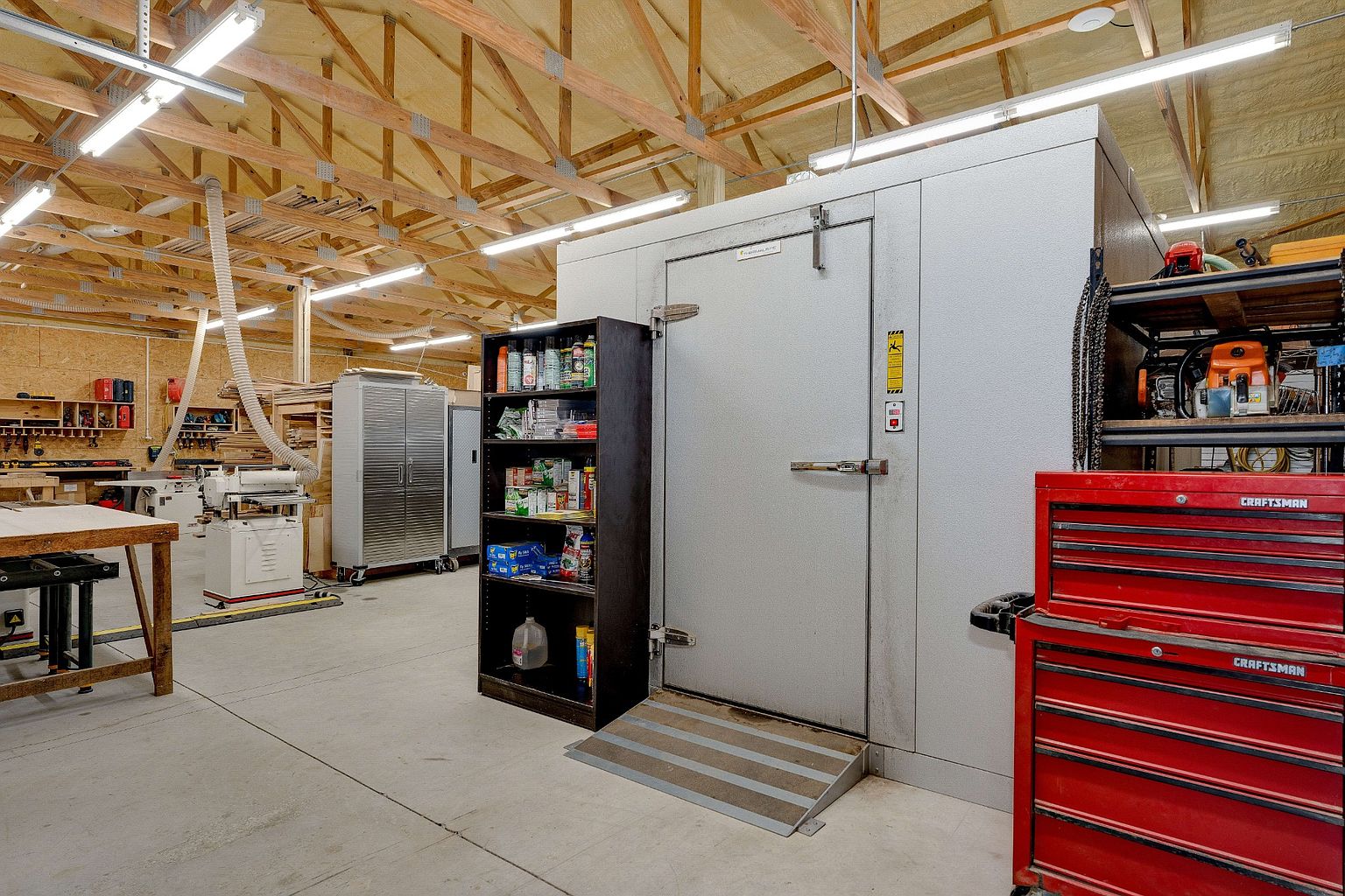 This image showcases a spacious workshop or garage interior, characterized by its high ceilings with exposed wooden beams and ample fluorescent lighting. The room features a large, walk-in cooler or storage unit, a red Craftsman tool chest, shelving units, and various woodworking tools and equipment. The concrete floor and organized layout suggest a functional and well-maintained workspace.
