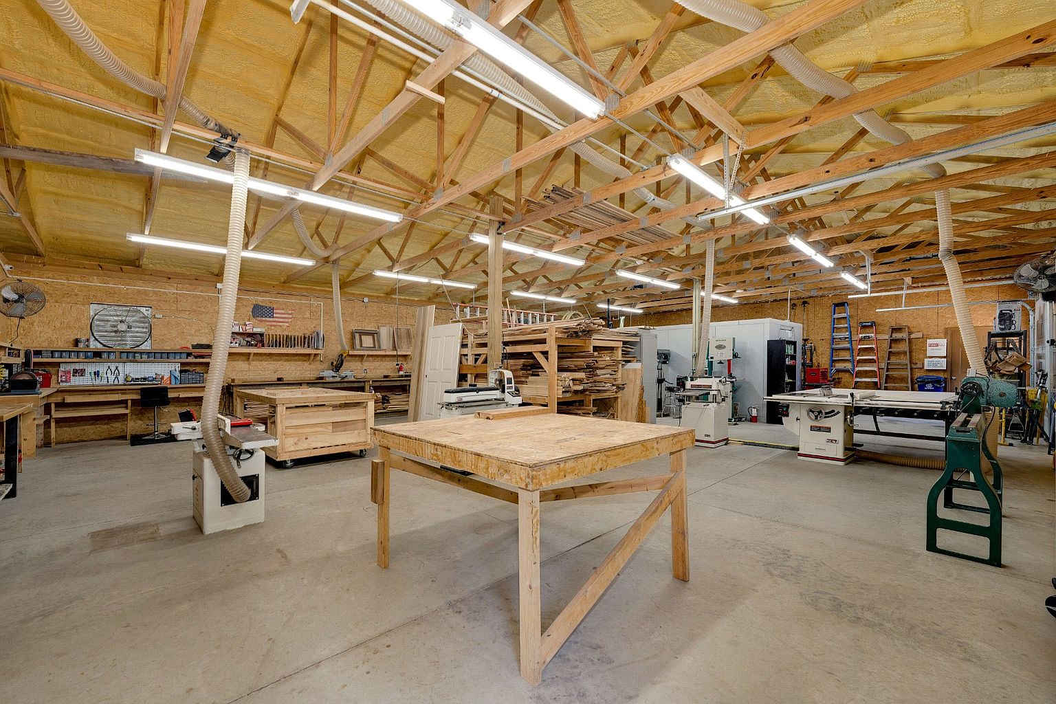 This is a spacious workshop featuring a high ceiling with exposed wooden beams and fluorescent lighting. The room is filled with various woodworking tools and equipment, including workbenches, saws, and storage racks filled with lumber. The concrete floor and ample space suggest a functional and well-equipped area for crafting and building.