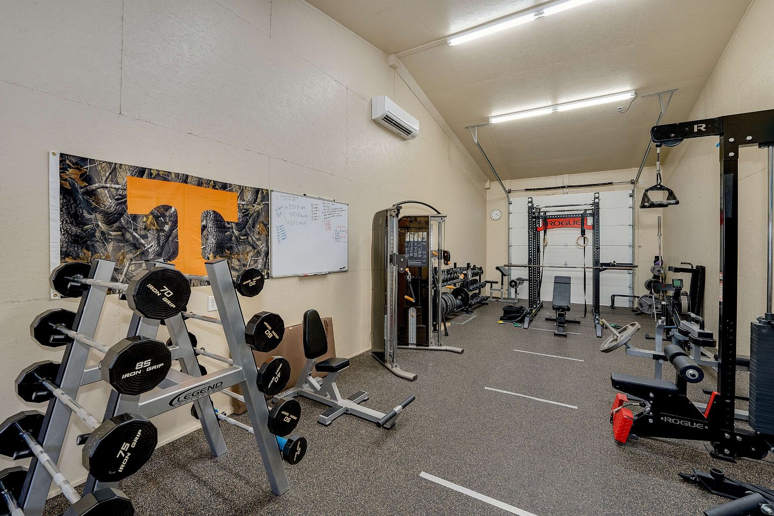 This is a well-equipped home gym featuring a variety of weightlifting equipment, including a weight rack with dumbbells, a cable machine, and a Rogue power rack. The floor is covered in a durable rubber material, and the walls are painted in a neutral tone. The space is well-lit with overhead fluorescent lights, creating a functional and motivating workout environment.